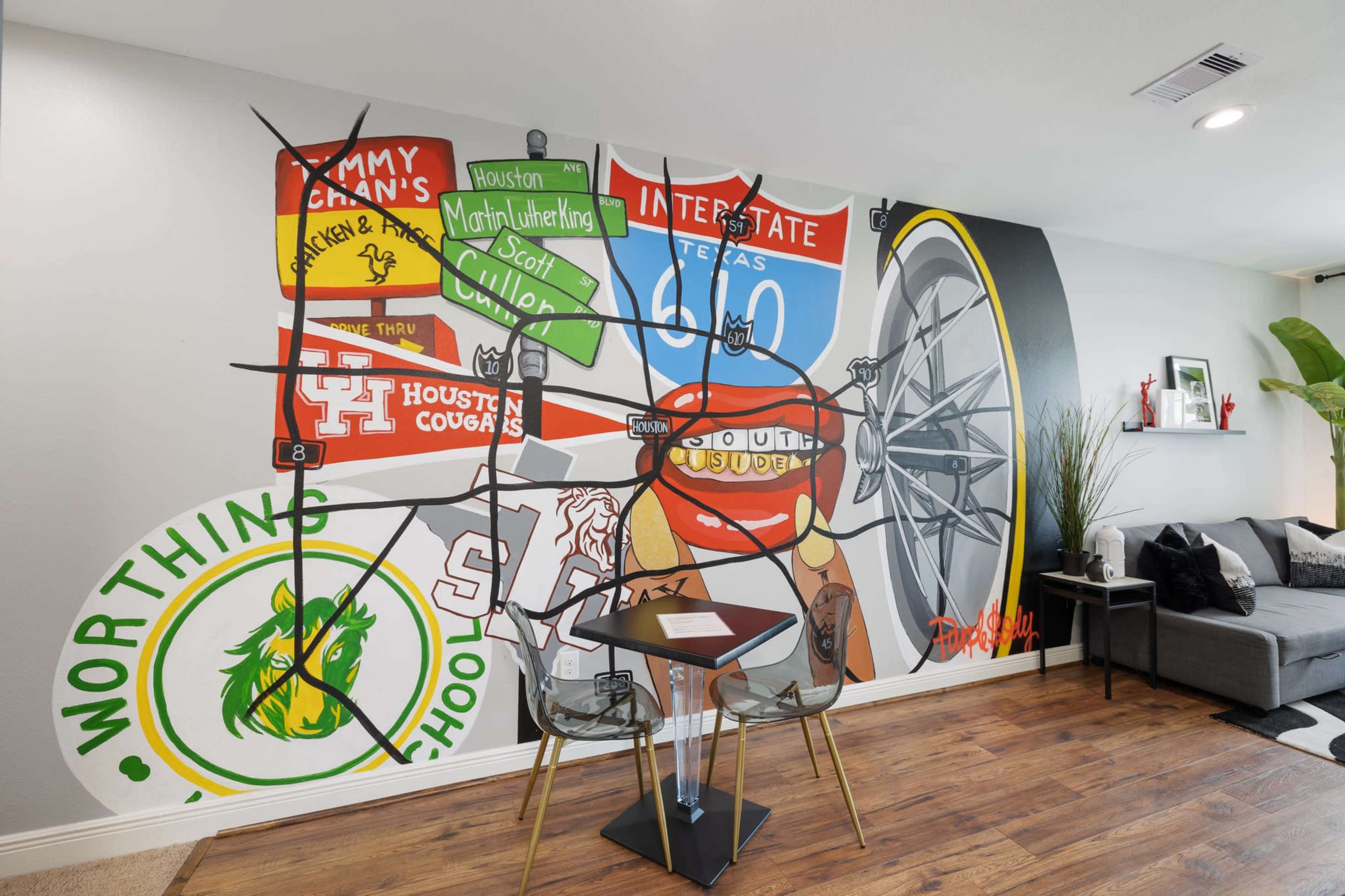 The image shows a modern living space featuring a colorful mural on the wall and a small black table with two clear chairs.