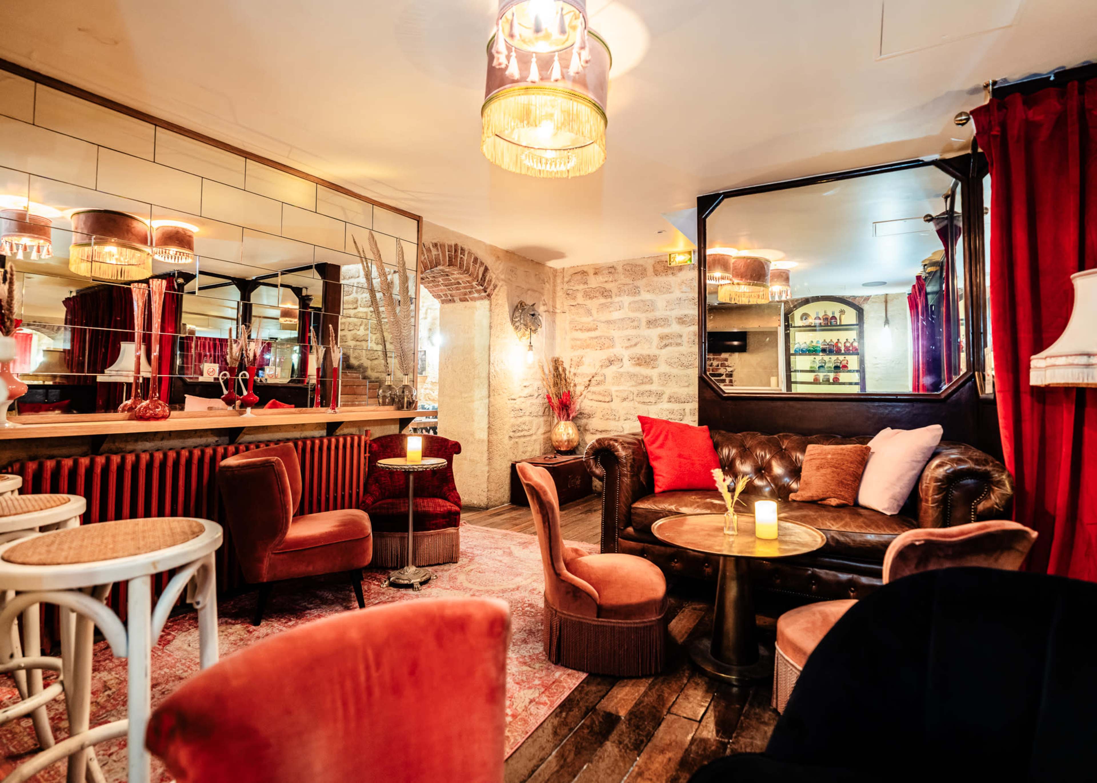 A stylish lounge area features a leather sofa, circular table, and illuminated chandeliers, with mirrors and red drapes adding to the ambiance.
