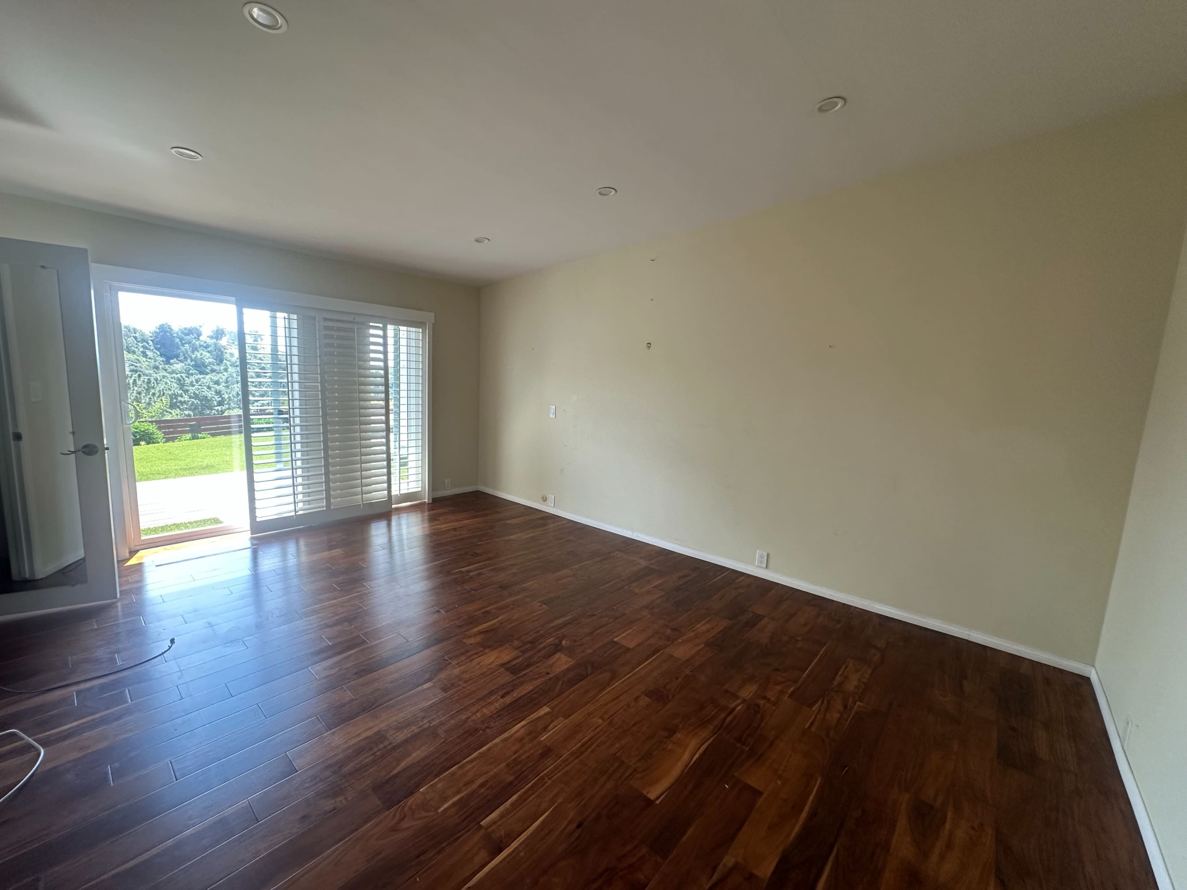 The image shows an empty room with wooden flooring, a sliding door leading to a patio, and shutters on the windows.