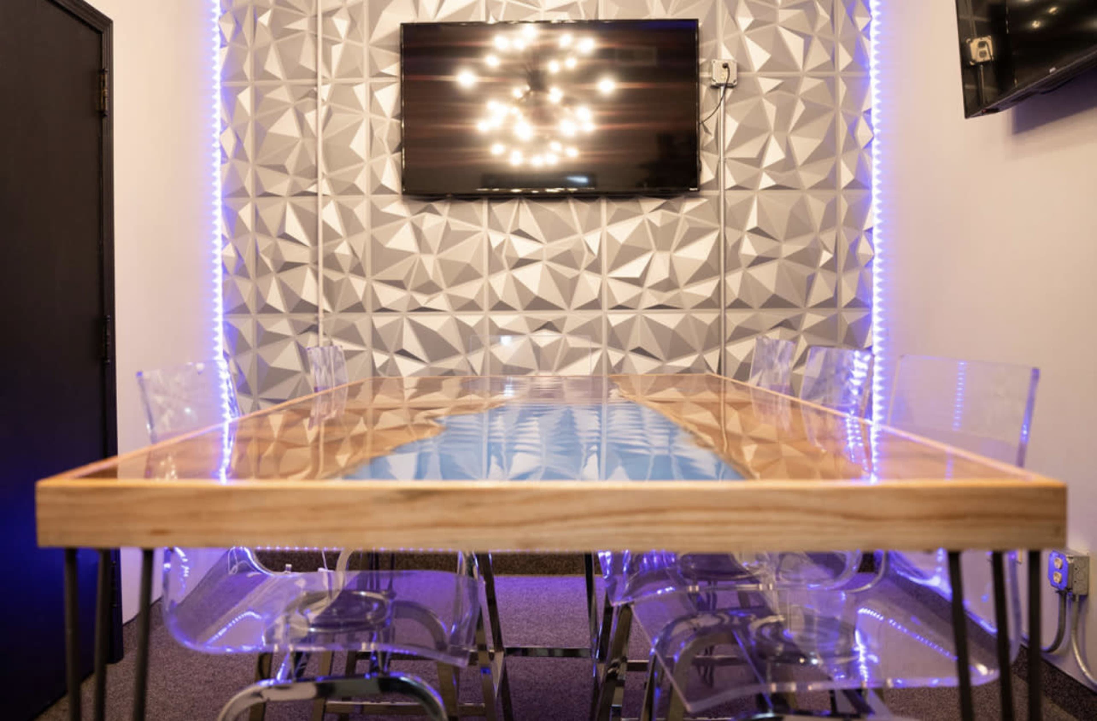 A modern conference room with a large wooden table and clear acrylic chairs, complemented by a geometric wall design and a wall-mounted television.