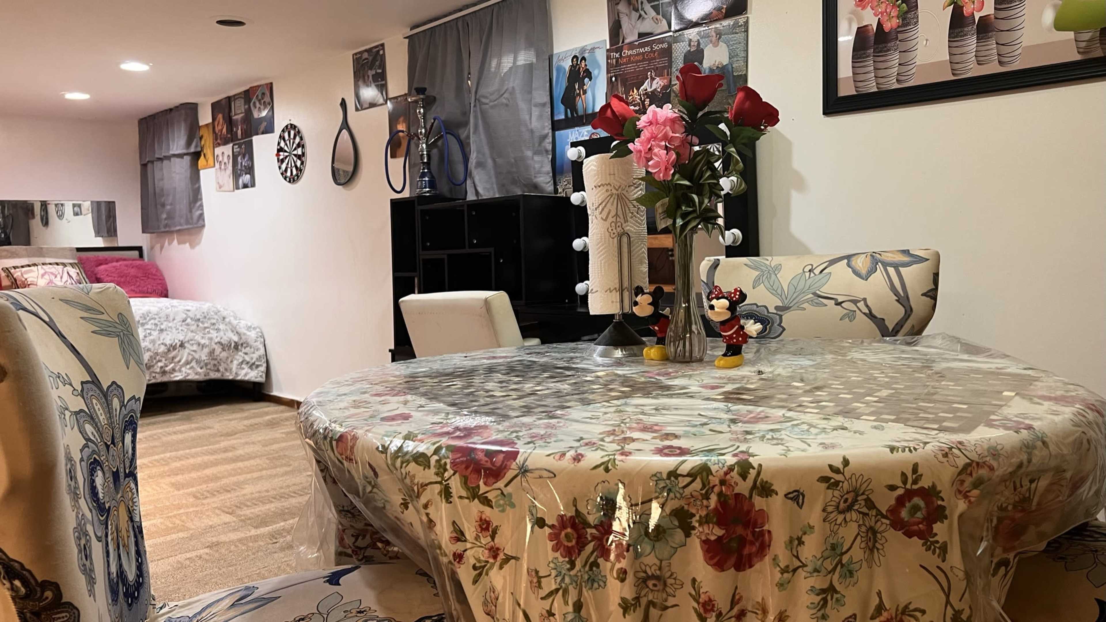 A cozy, semi-furnished room with a dining table set with a floral tablecloth, a vase of roses, and decorative items, alongside a bed in the background and posters on the walls.