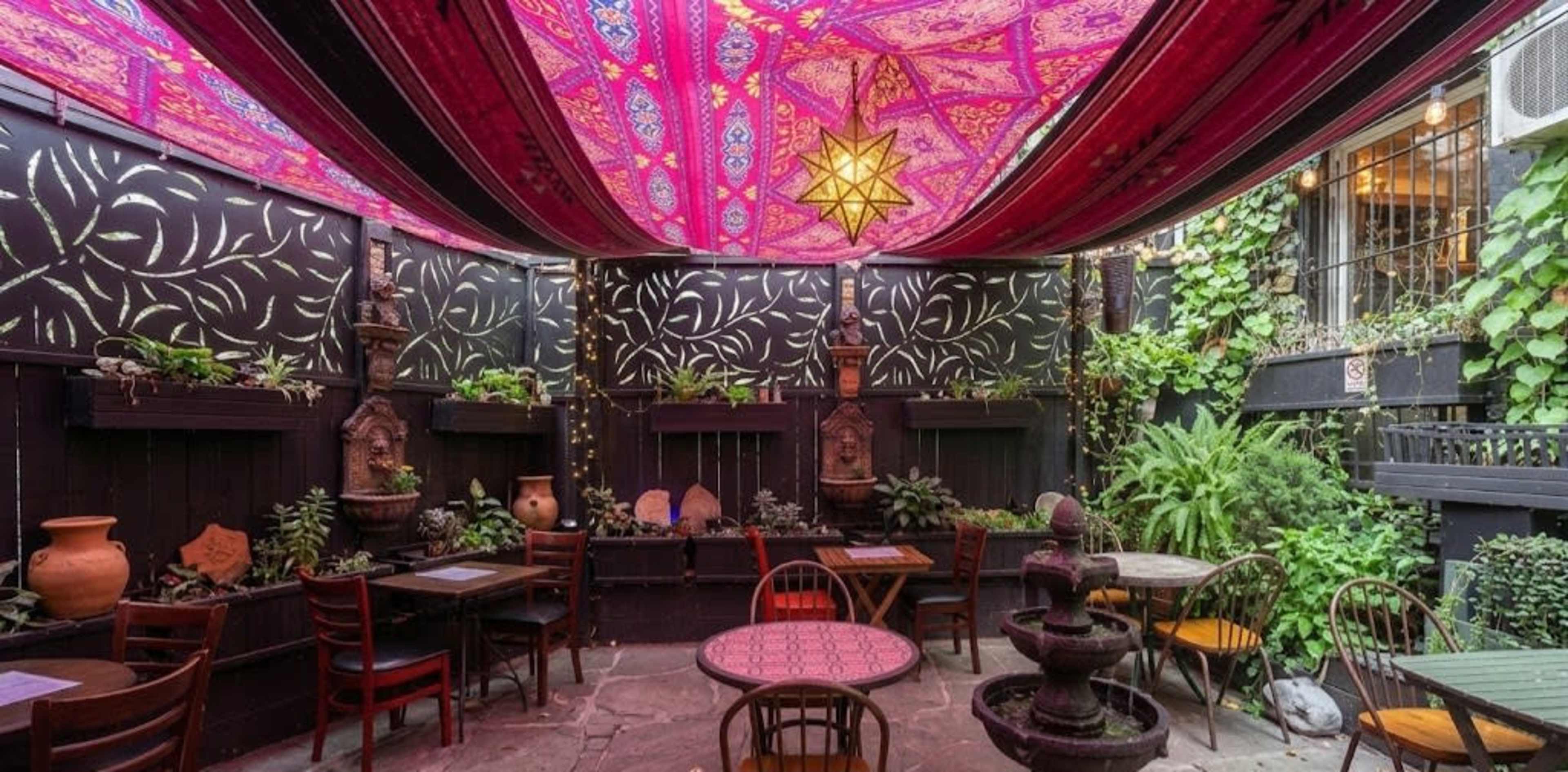 The image shows a cozy outdoor dining area decorated with vibrant fabric overhead, surrounded by greenery and featuring small fountains and tables.