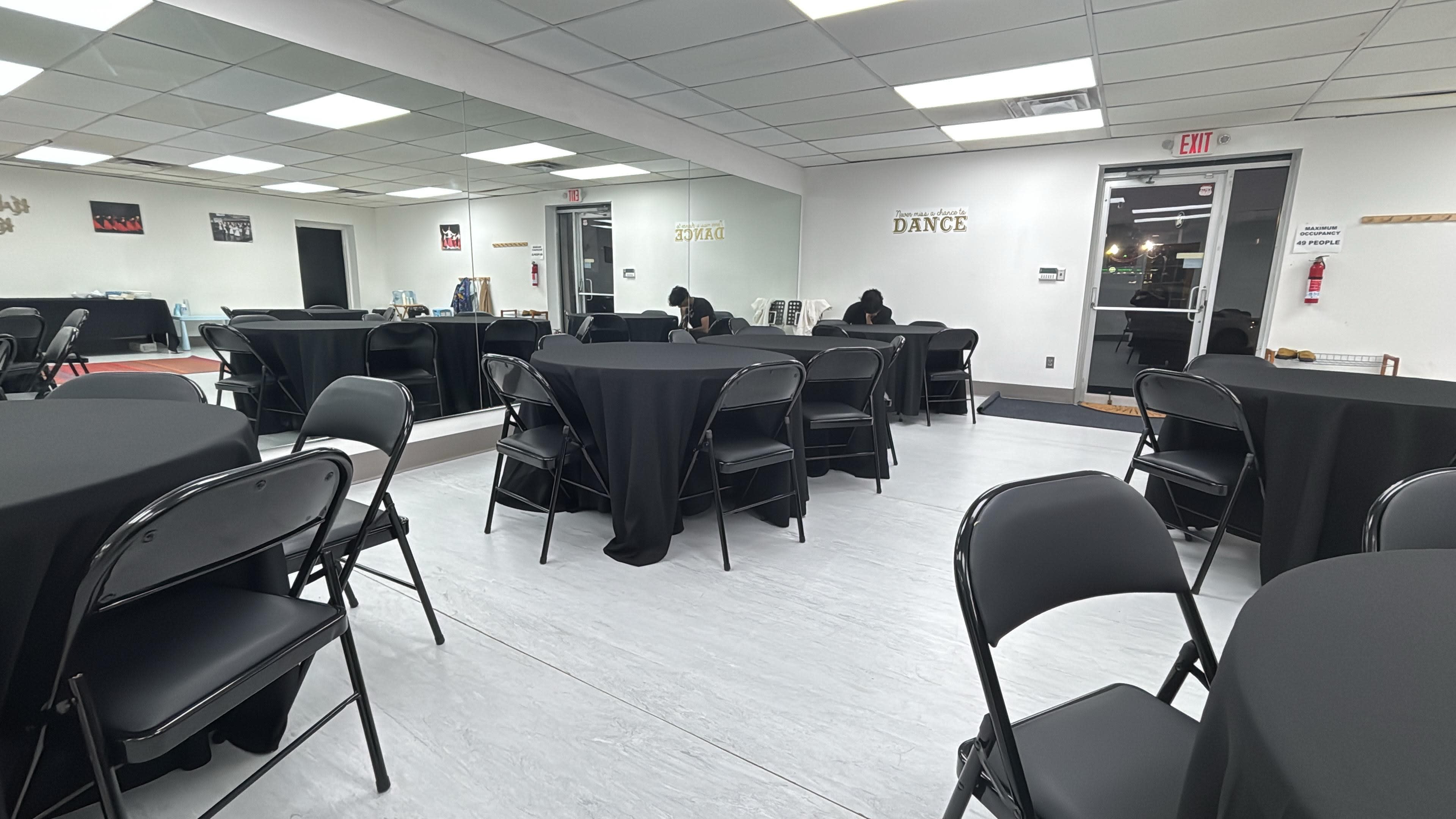 The room features black tables and chairs arranged for an event, with mirrors on one wall and several people seated at the tables.