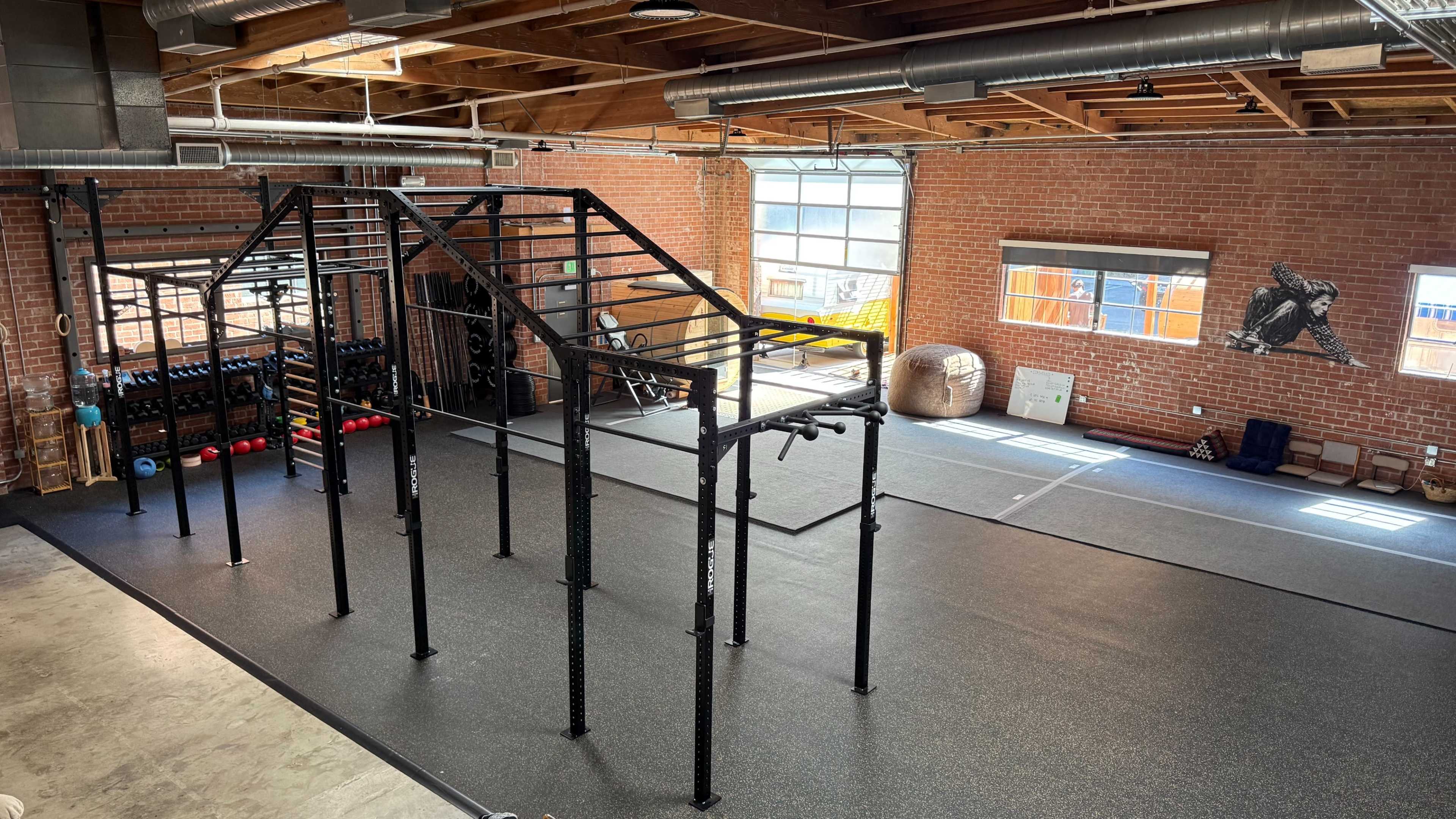 The image shows a spacious gym with large windows, featuring various exercise equipment, a rig for workouts, and a rubber flooring surface.