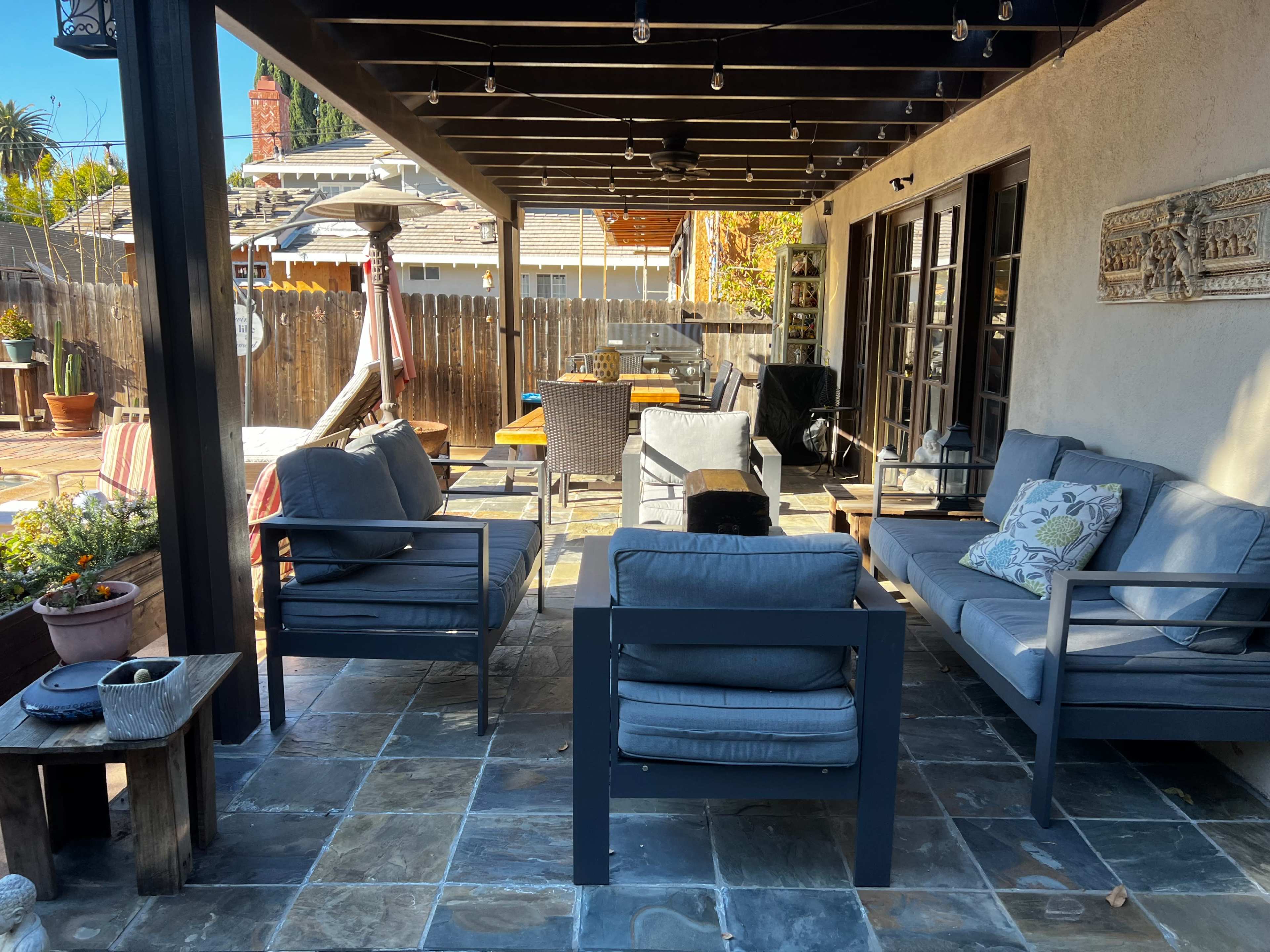 A patio area furnished with gray seating, a table, and various potted plants, surrounded by a wooden fence and outdoor decorations.