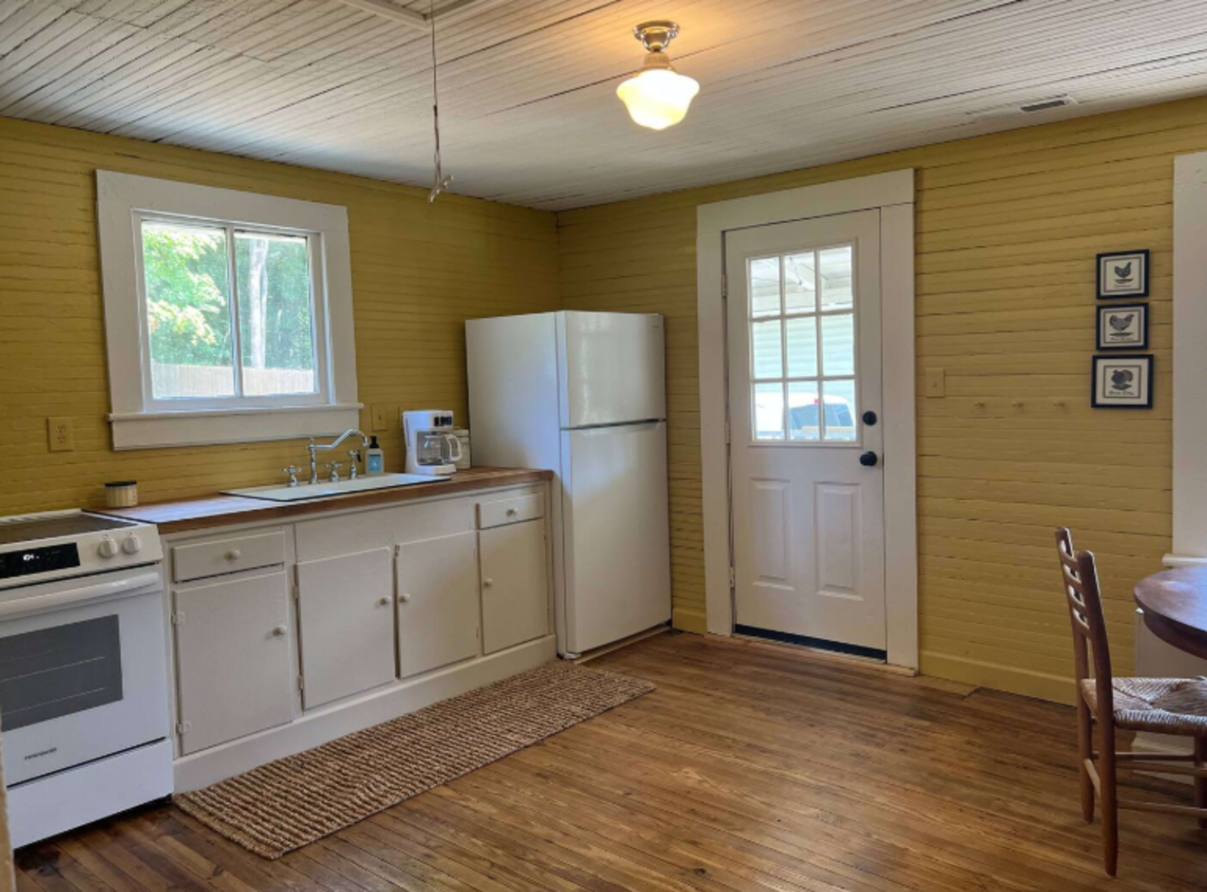 The image shows a bright yellow kitchen featuring a white refrigerator, a stove, a sink area with counter space, and a wooden dining table near a door leading outside.