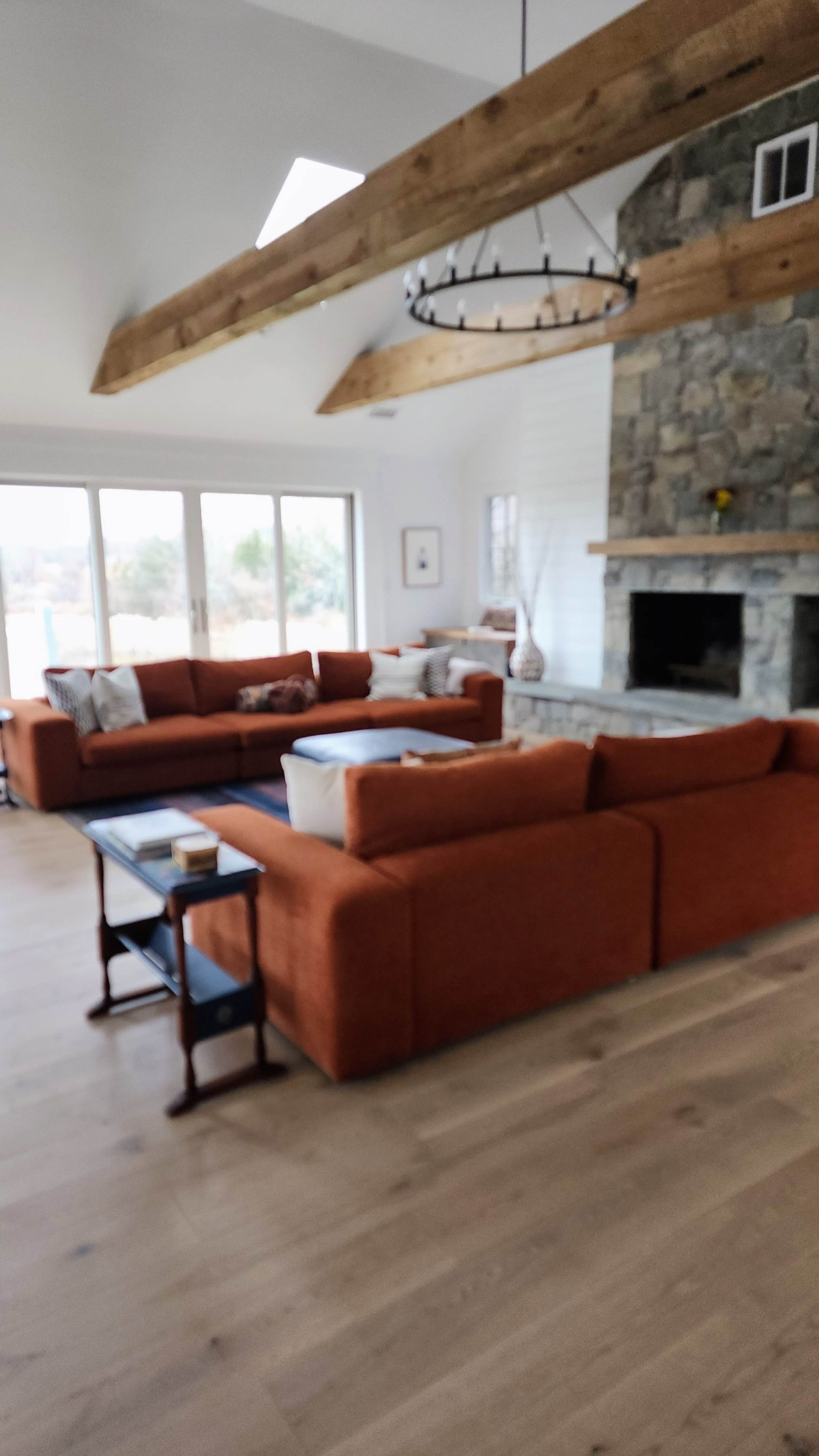 The image shows a spacious living room featuring a stone fireplace, orange sectional sofas, large windows, and wooden beams overhead.