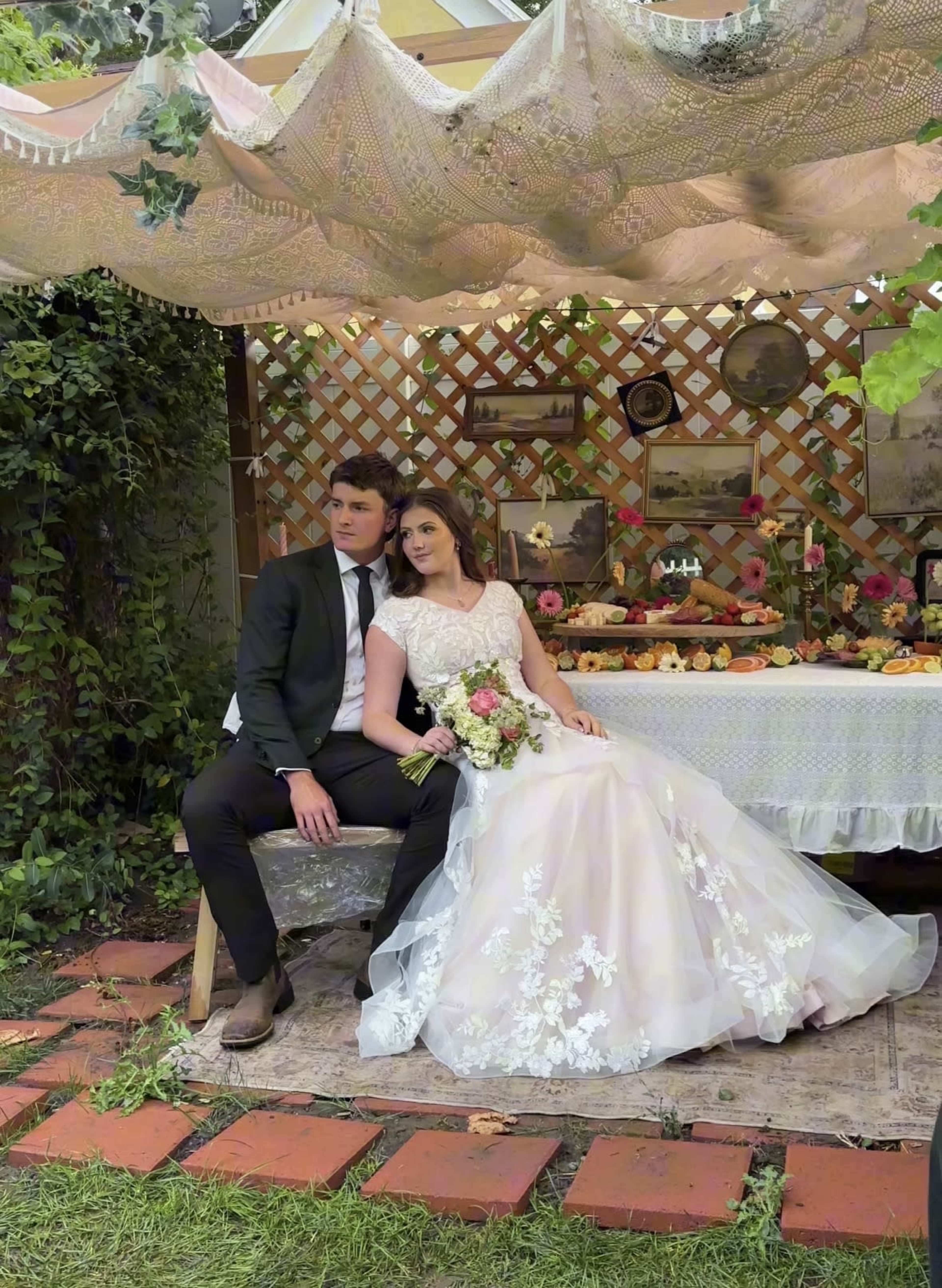 A couple poses for a photo in a garden setting, sitting against a backdrop adorned with flowers and vintage decor.