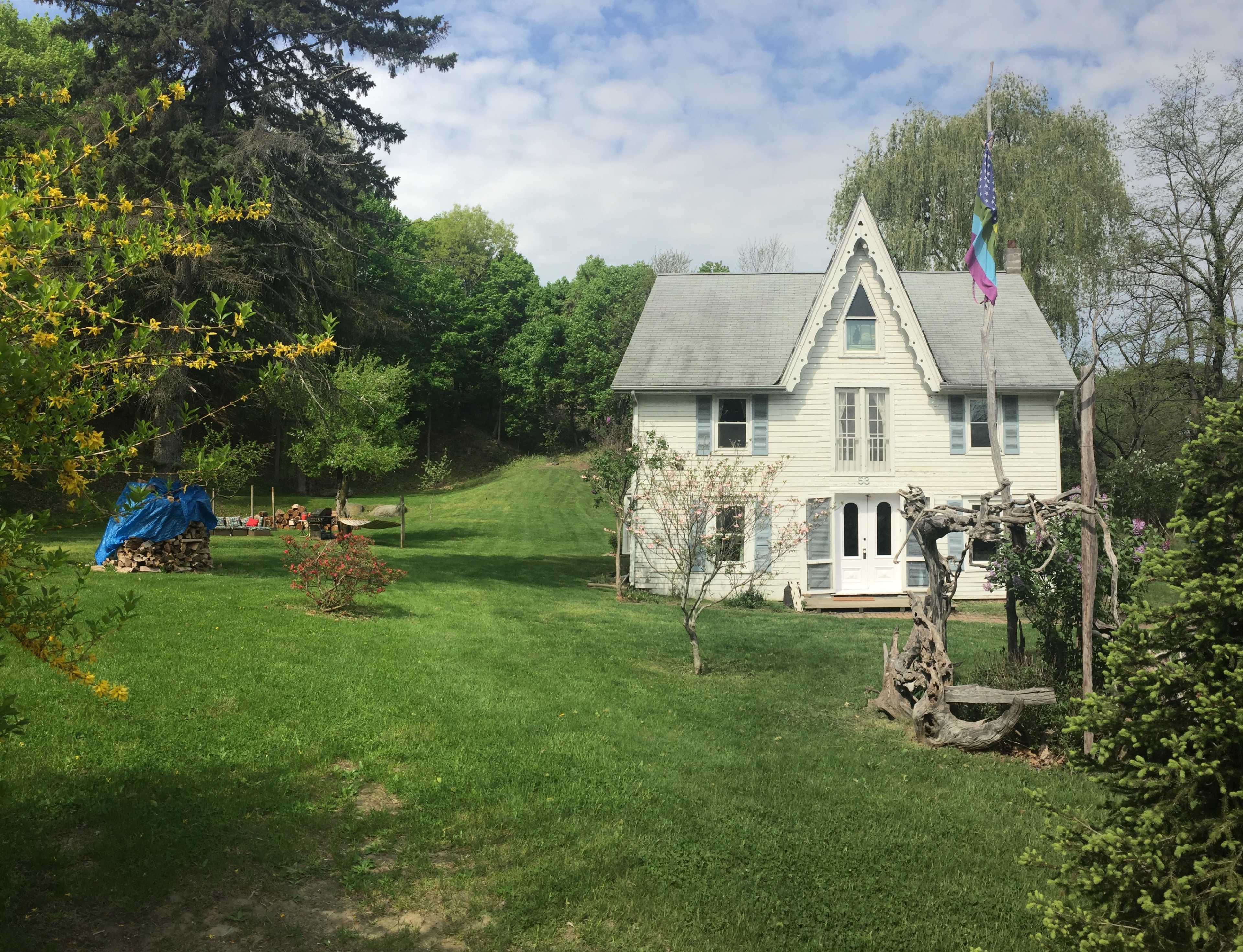 A two-story house with a pointed roof and flags stands in a green yard surrounded by trees and a wooden swing set.
