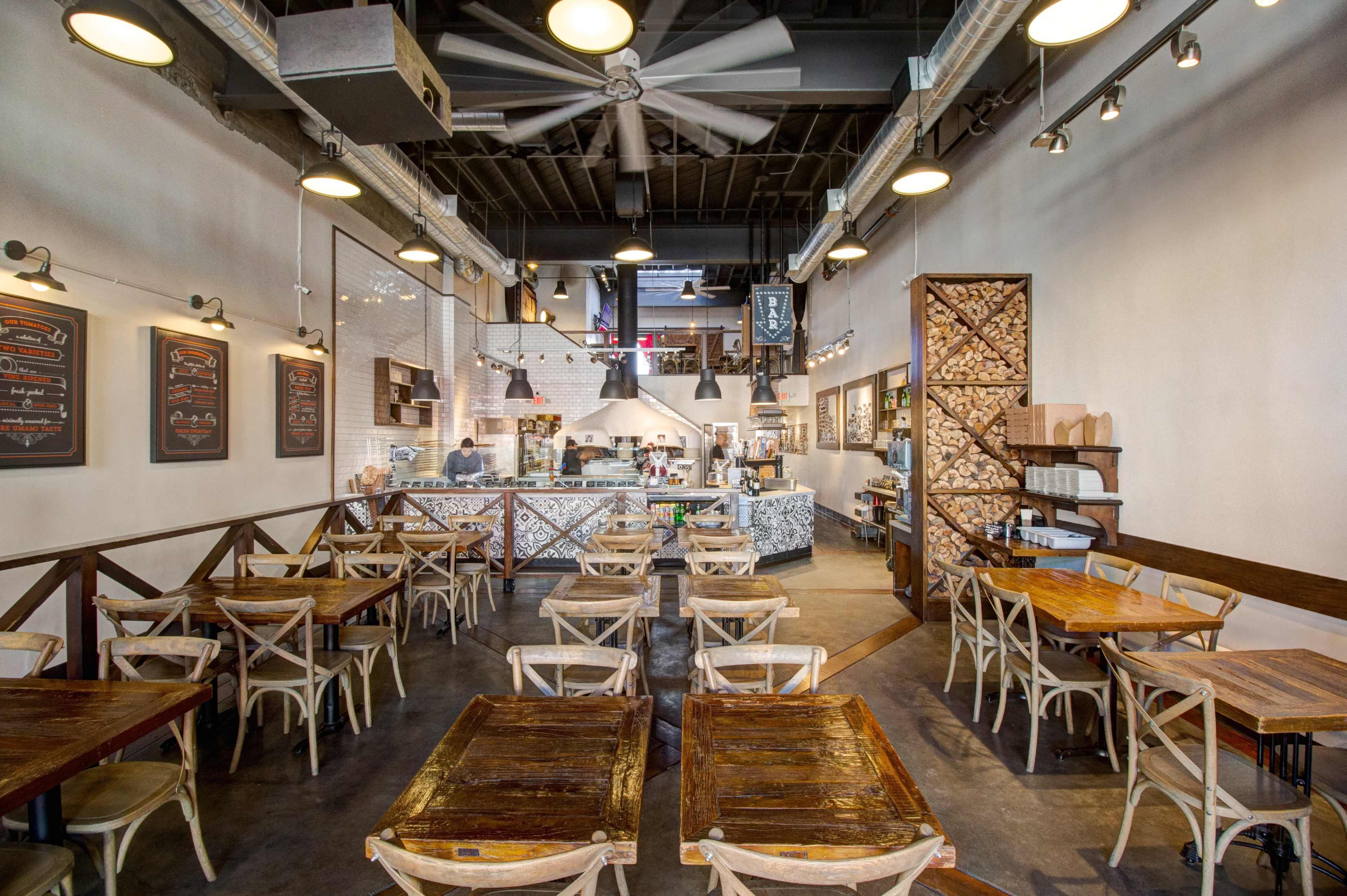 A restaurant interior with wooden tables and chairs arranged neatly in an open space, facing a counter where food is prepared.