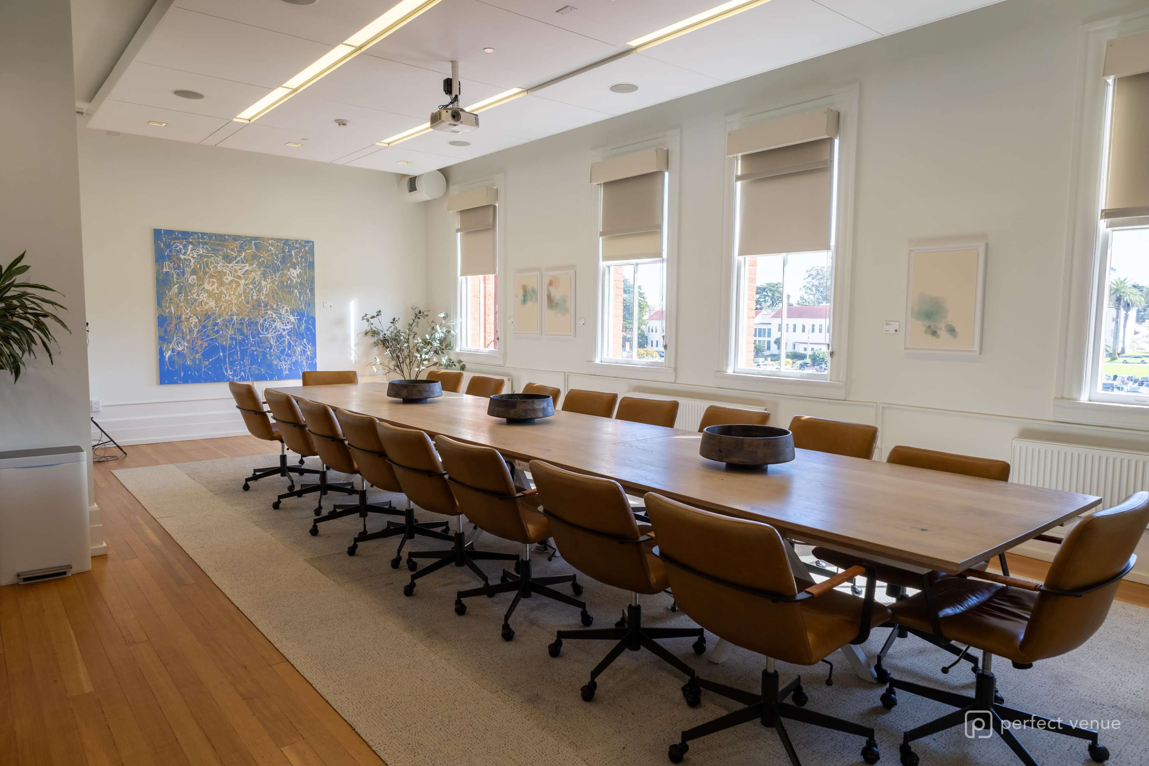 The image shows a spacious conference room with a long wooden table surrounded by twelve leather chairs, large windows, and abstract artwork on the walls.