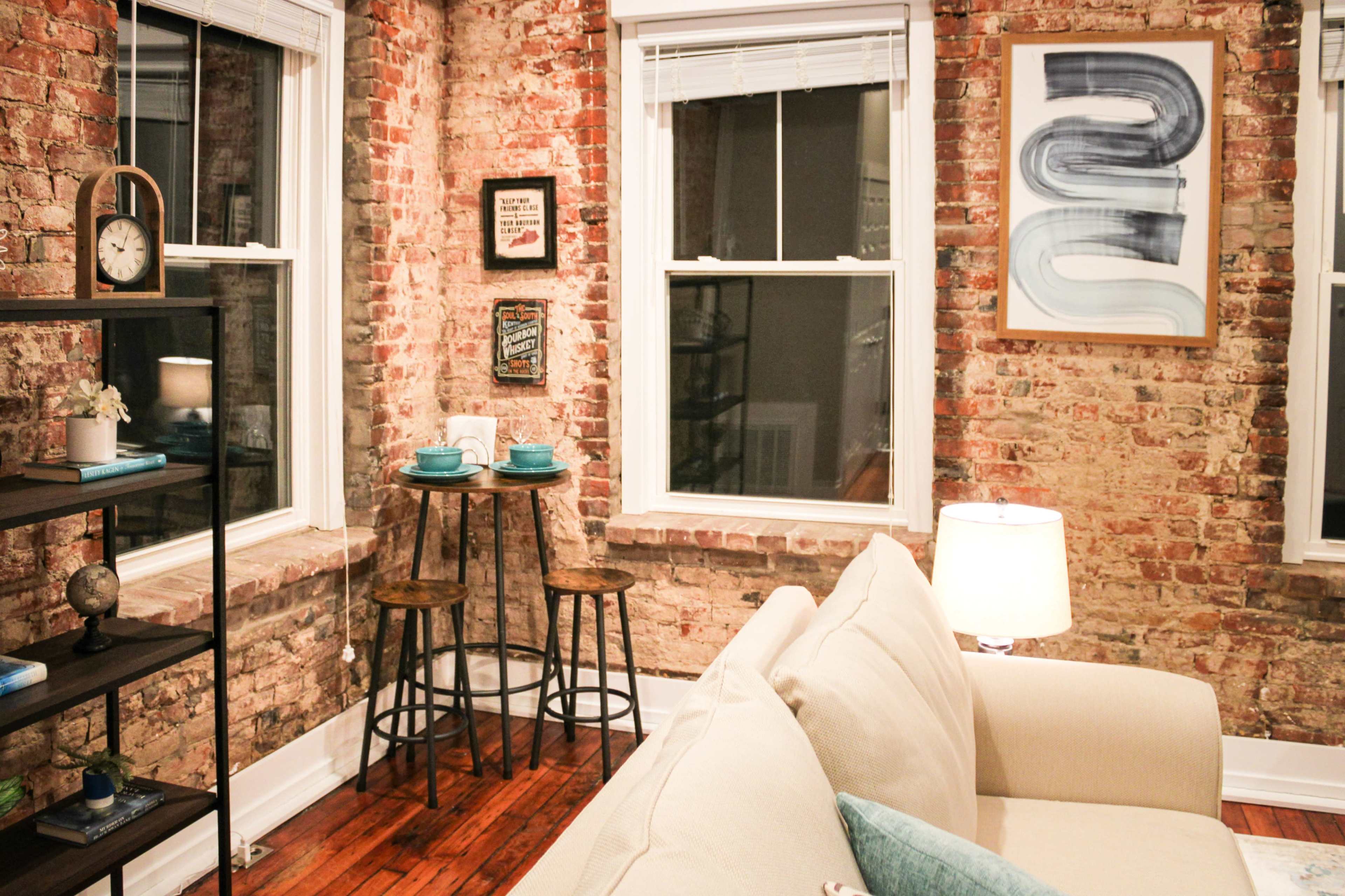 A cozy living area with exposed brick walls, two stools under a small table, and a sofa facing the windows.