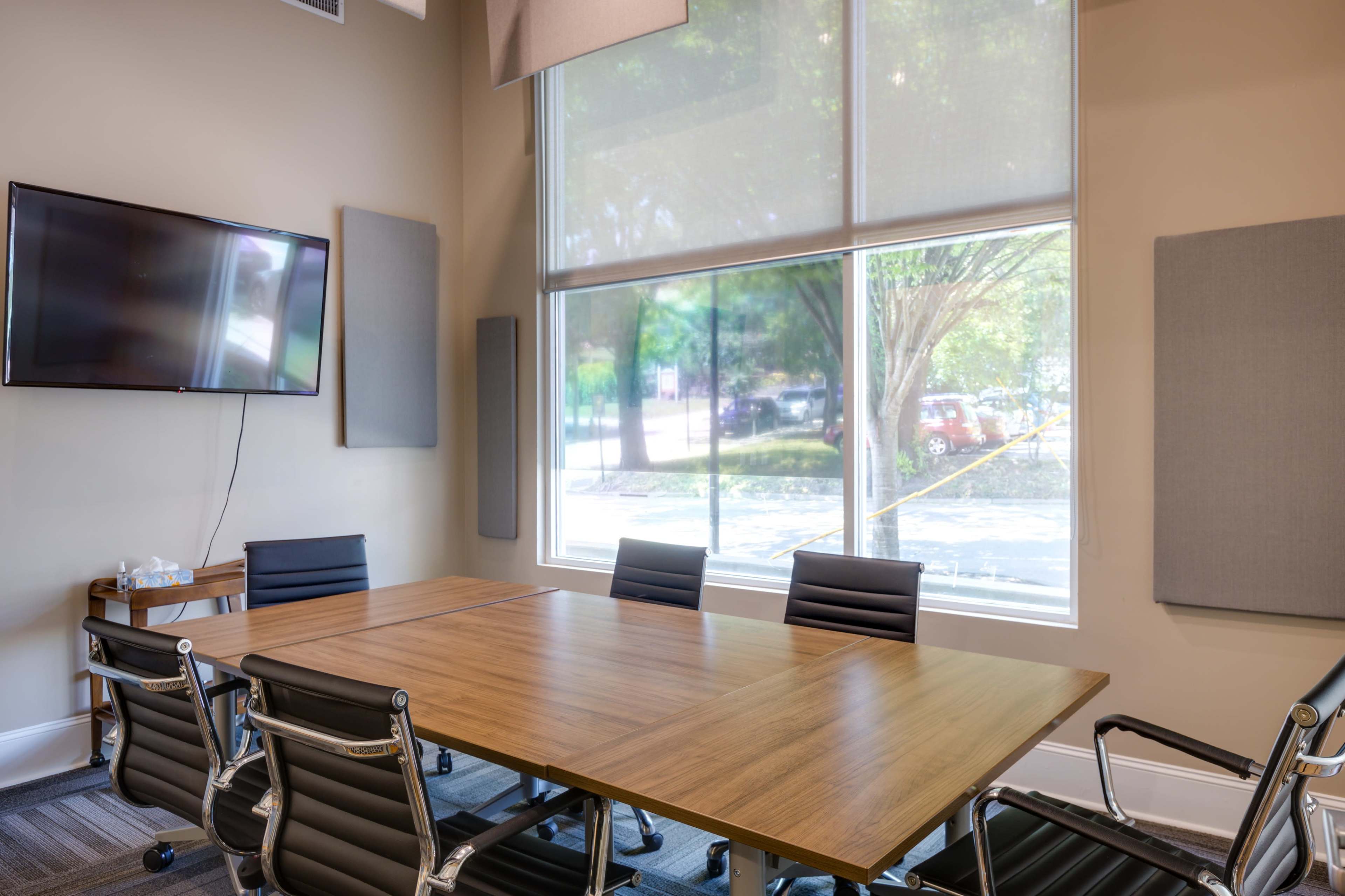 The image shows a modern conference room with a large wooden table, black leather chairs, and a television mounted on the wall, illuminated by natural light from large windows.