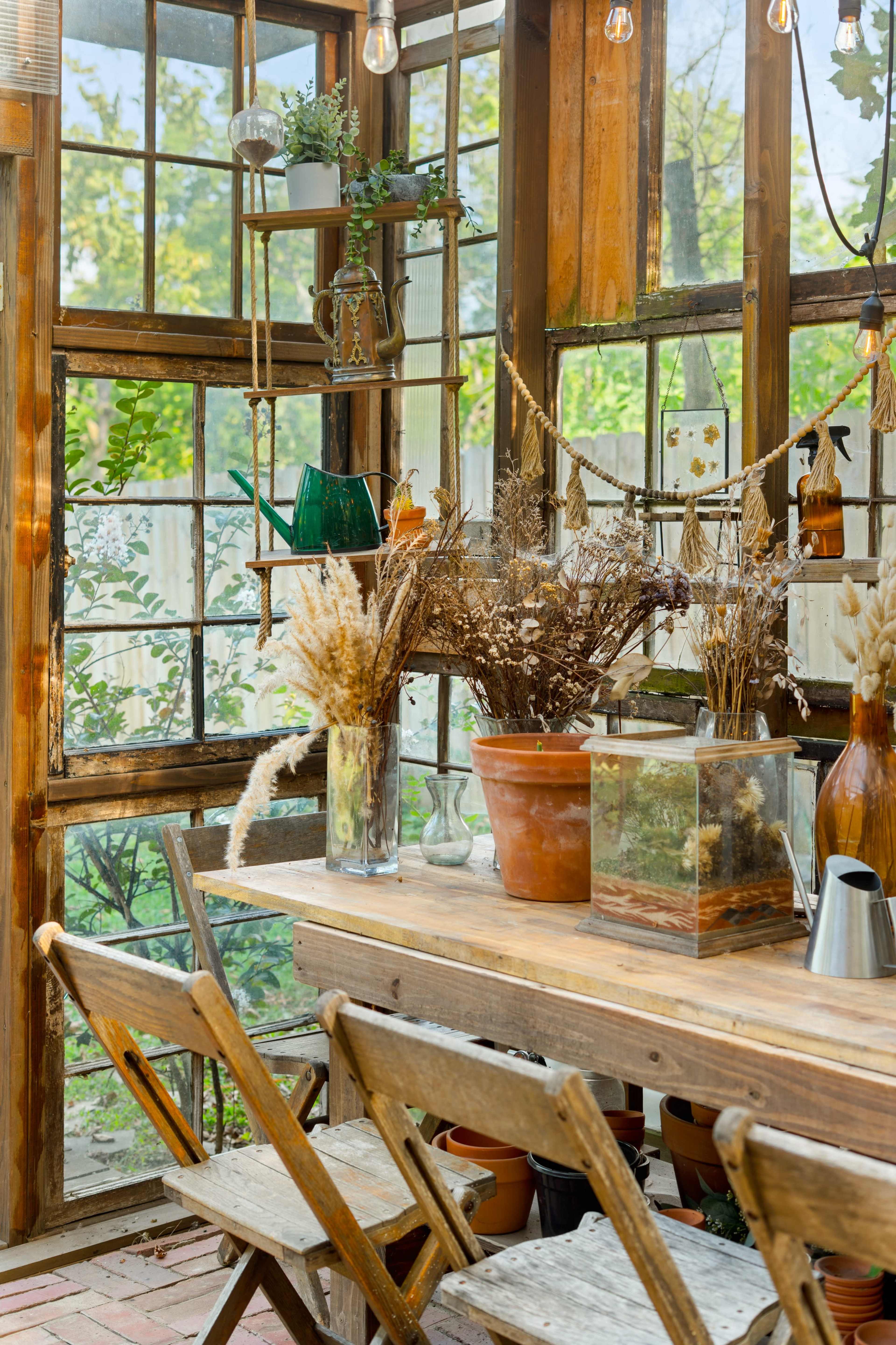 The image shows a cozy greenhouse interior with a wooden table surrounded by chairs, decorated with potted plants, dried flowers, and various glass containers.