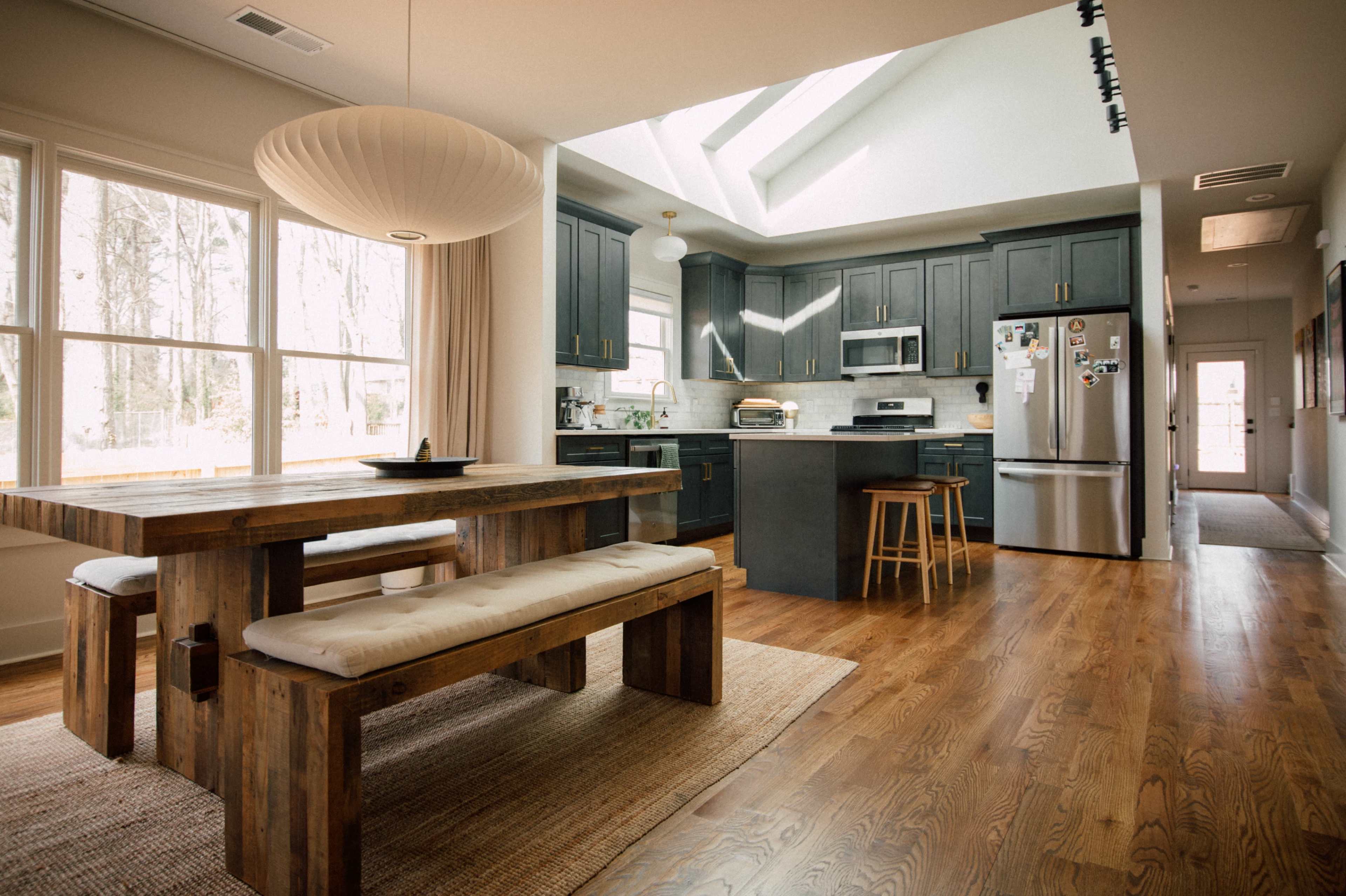 A modern kitchen features dark cabinetry, stainless steel appliances, and a wooden dining table with benches, illuminated by natural light through large windows.