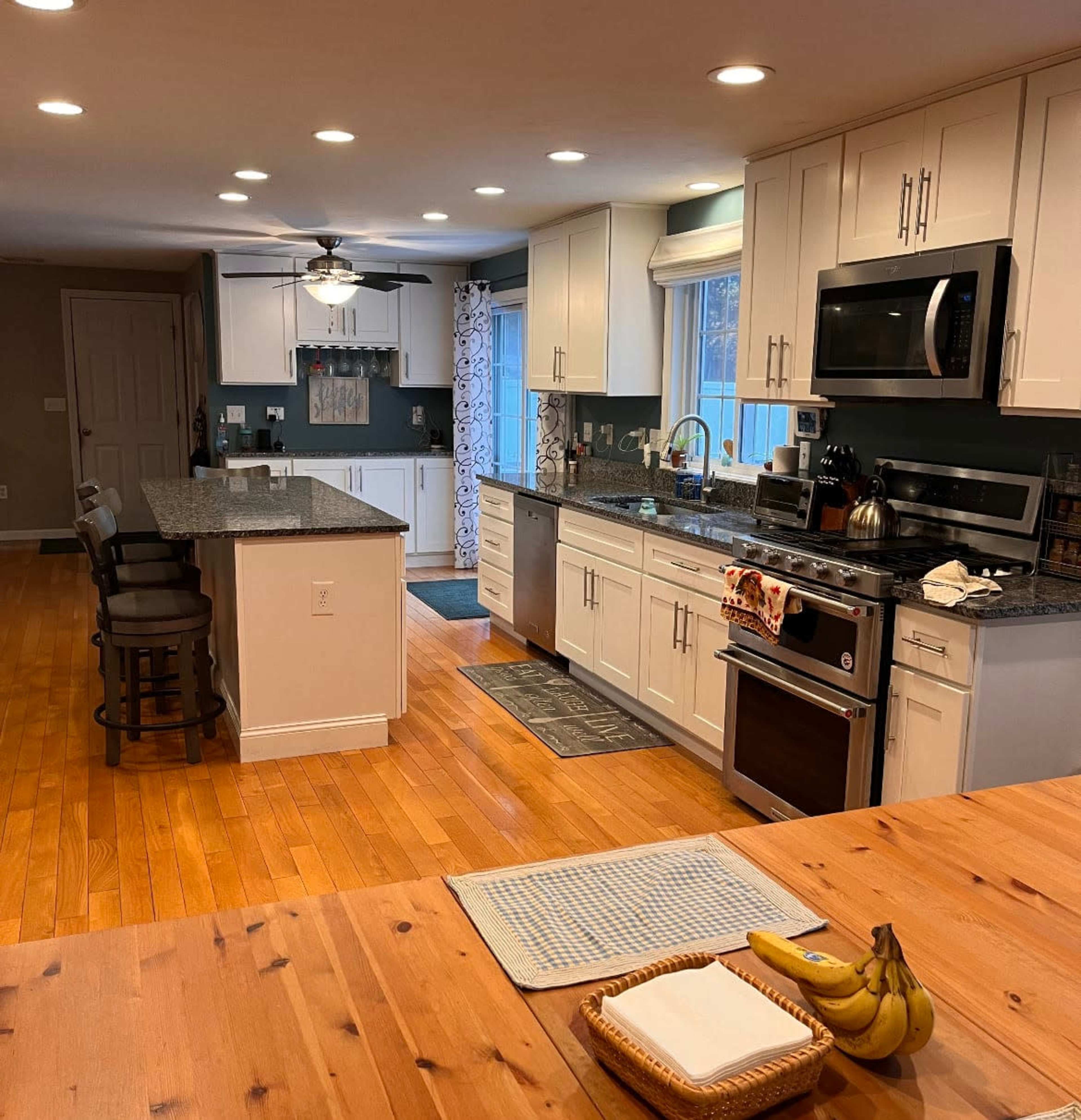 The image shows a modern kitchen with white cabinets, granite countertops, and stainless steel appliances, featuring a large island with bar stools and a wooden dining table with bananas on it.