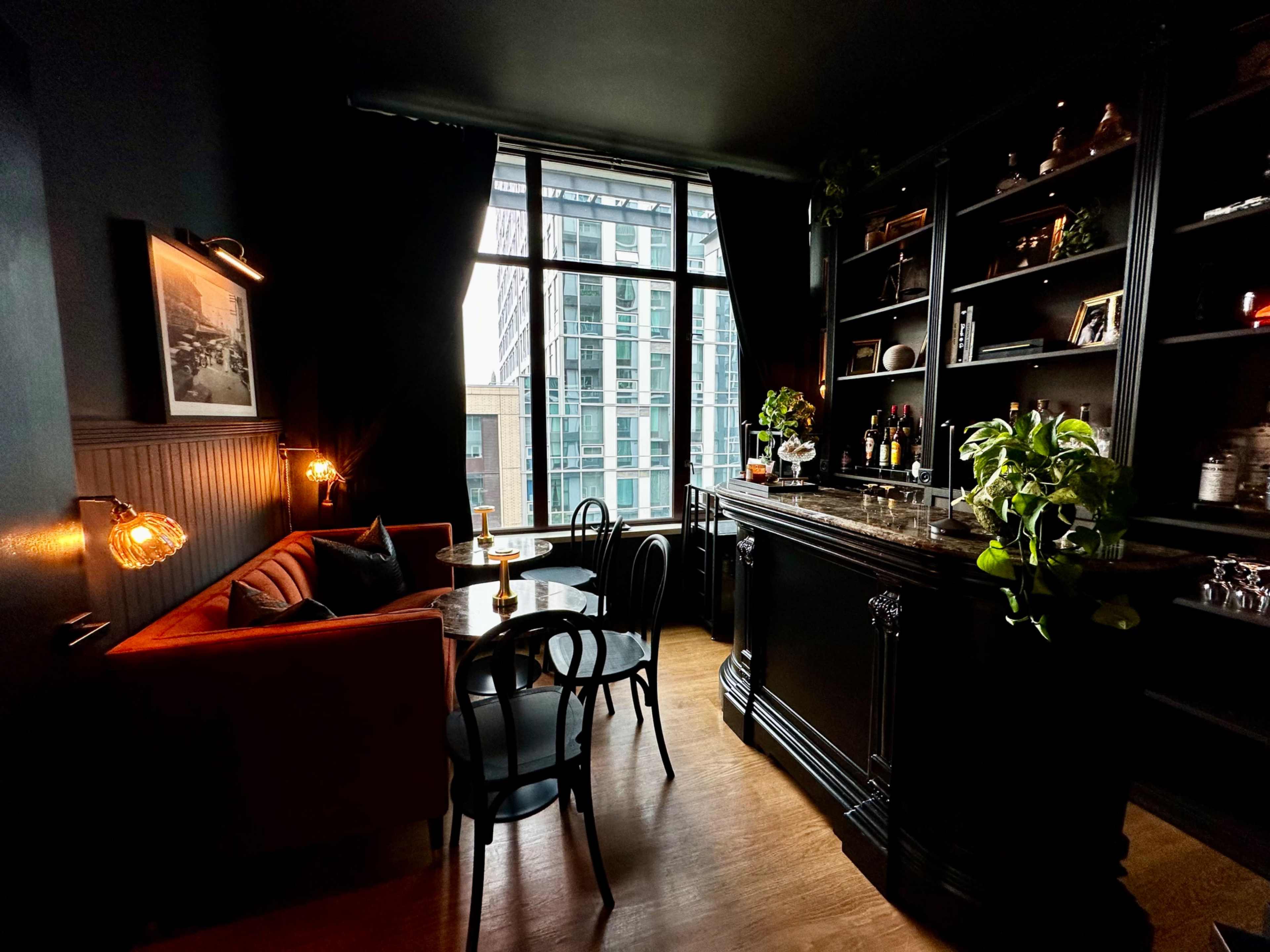 A cozy seating area with a dark-toned bar and large window, showcasing a modern urban view.