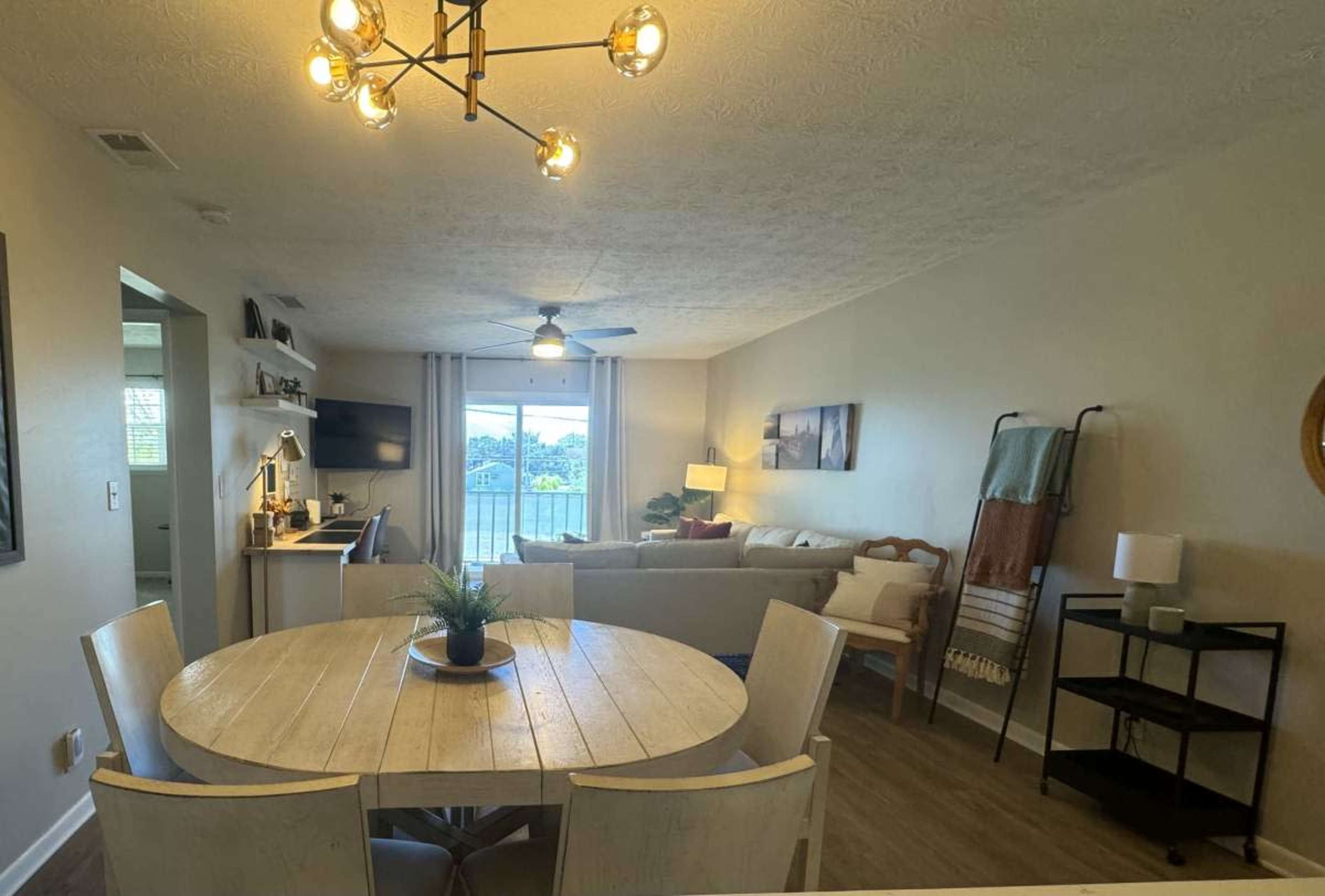 The image shows a modern living space featuring a round dining table, a sofa area with a television, and a balcony visible through large windows.