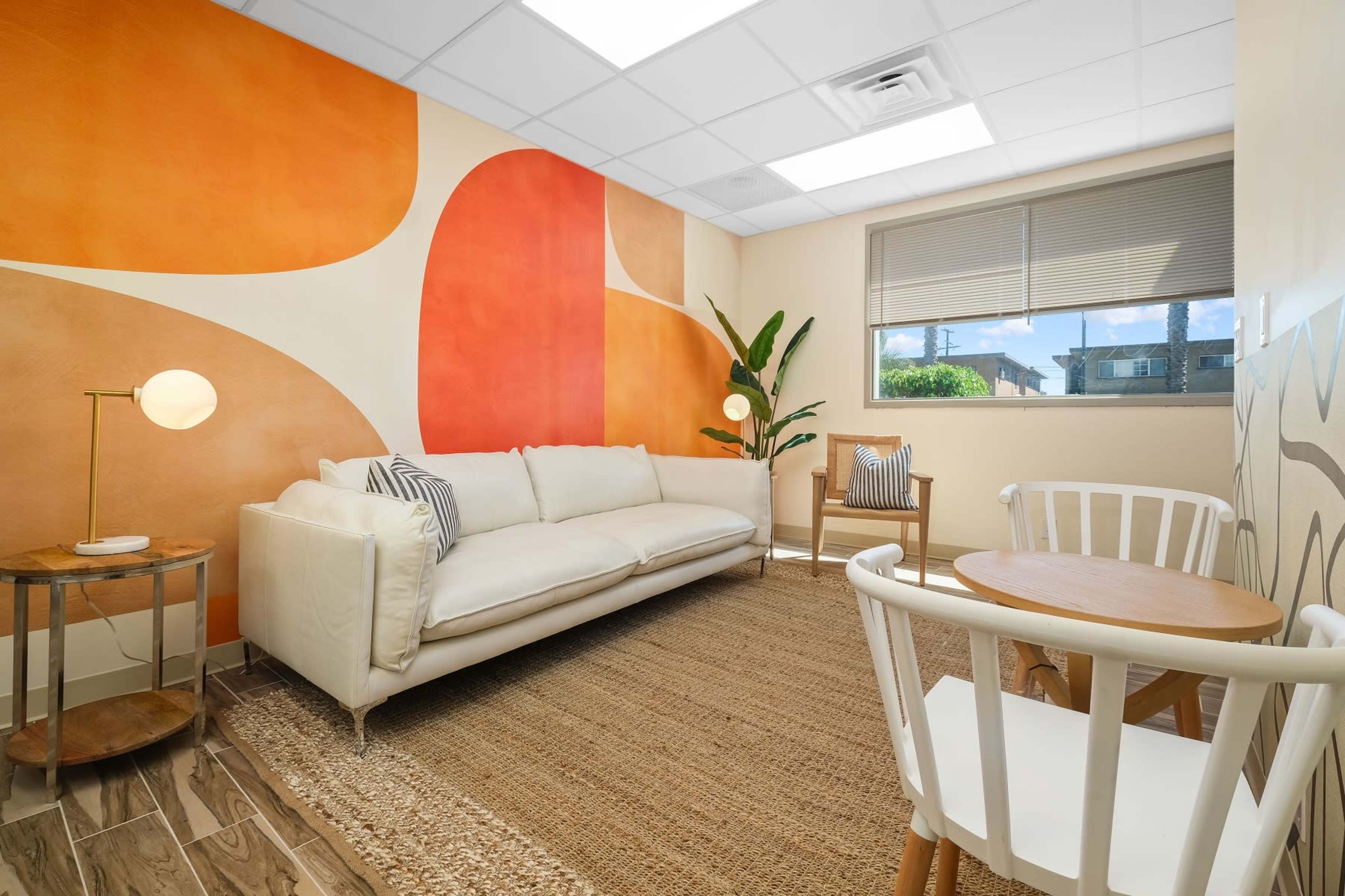 A well-lit room featuring a white sofa, a wooden chair, a small table, and a large wall mural with orange and beige patterns.