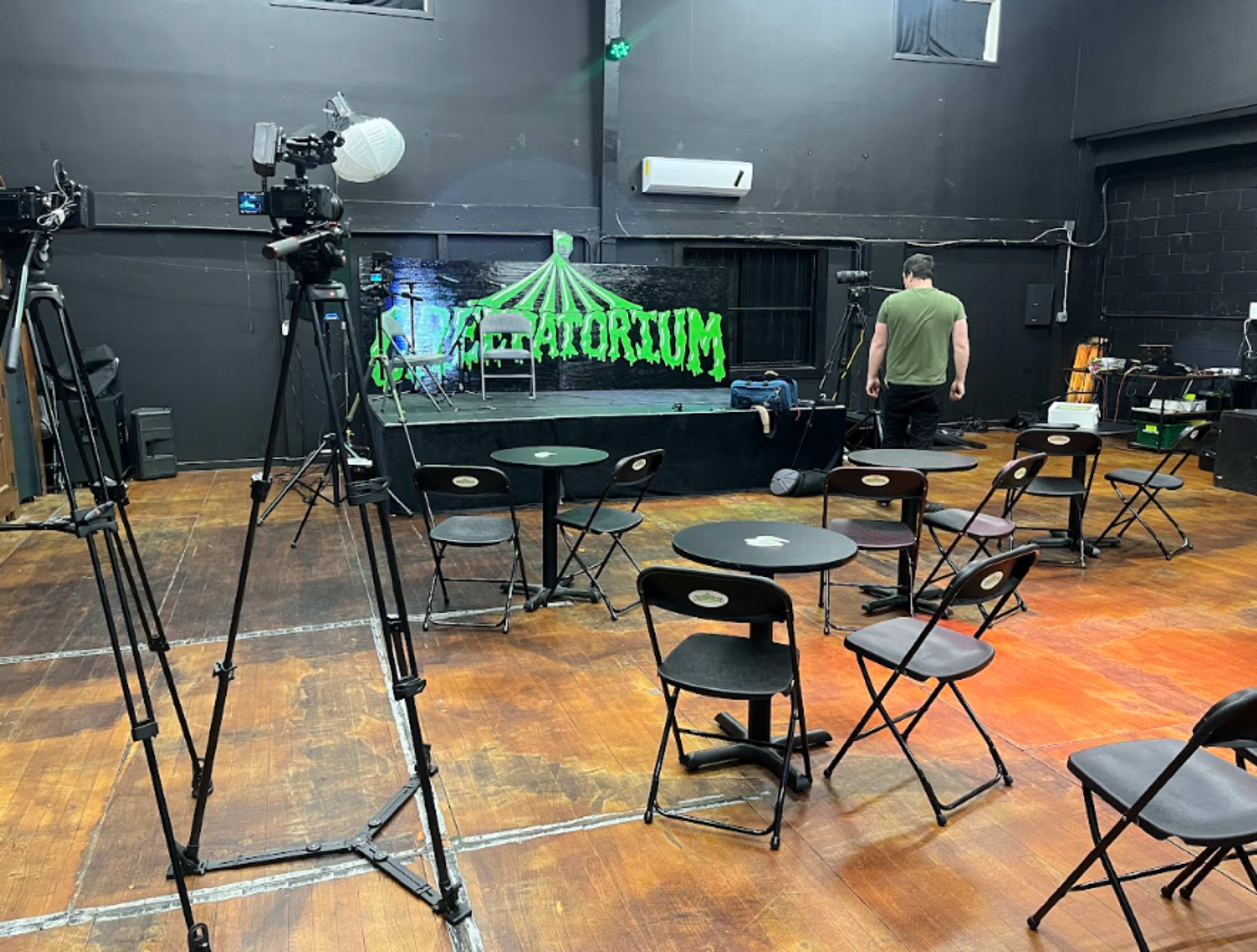 A stage is set up in a dark room with chairs arranged in front for an event, featuring green decorations and recording equipment.