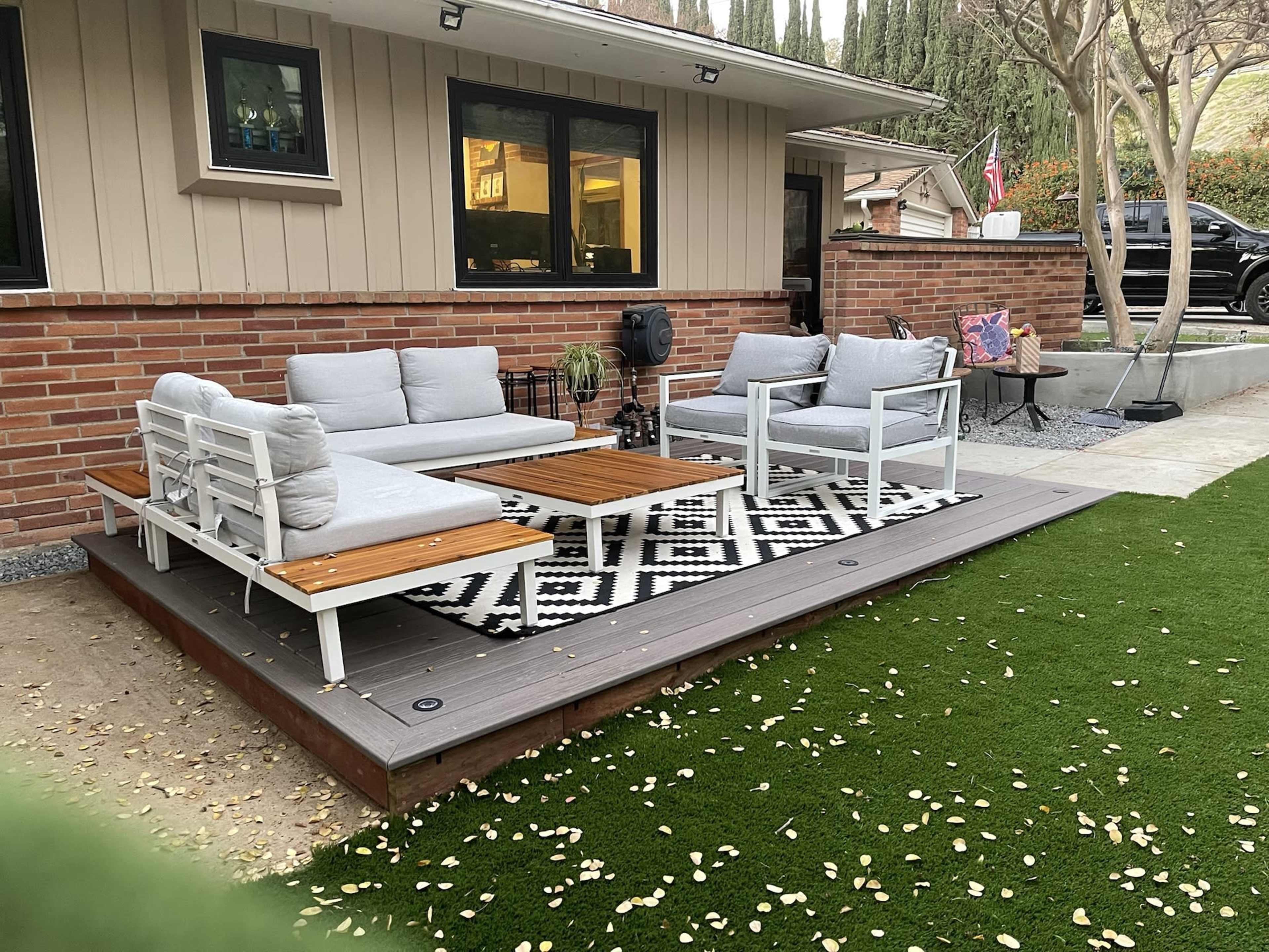 A outdoor seating area features a wooden deck with gray couches and a patterned rug surrounded by grass and landscaping.