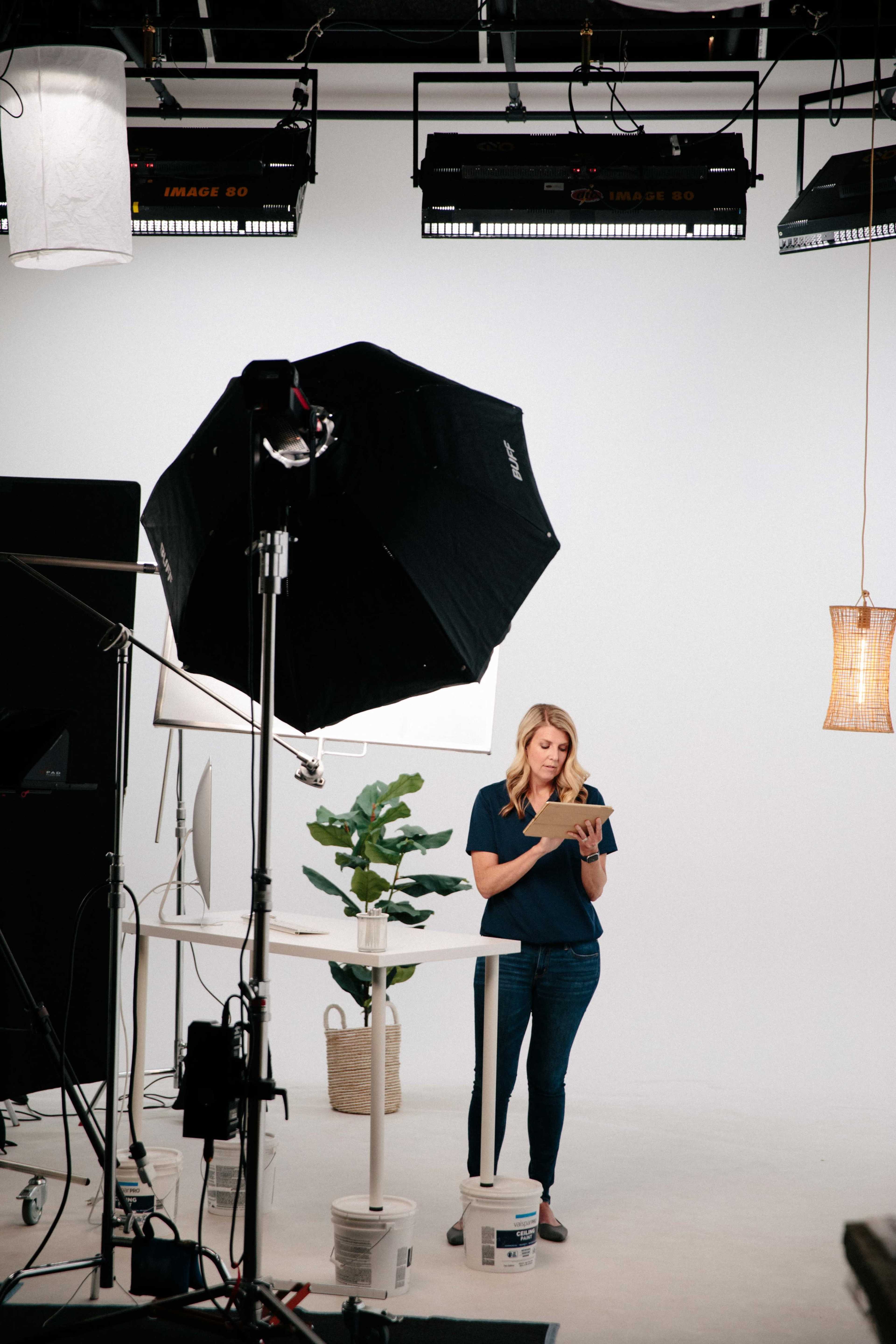 A woman stands near a table with a plant, holding a tablet in a well-lit studio setting equipped for a photoshoot.