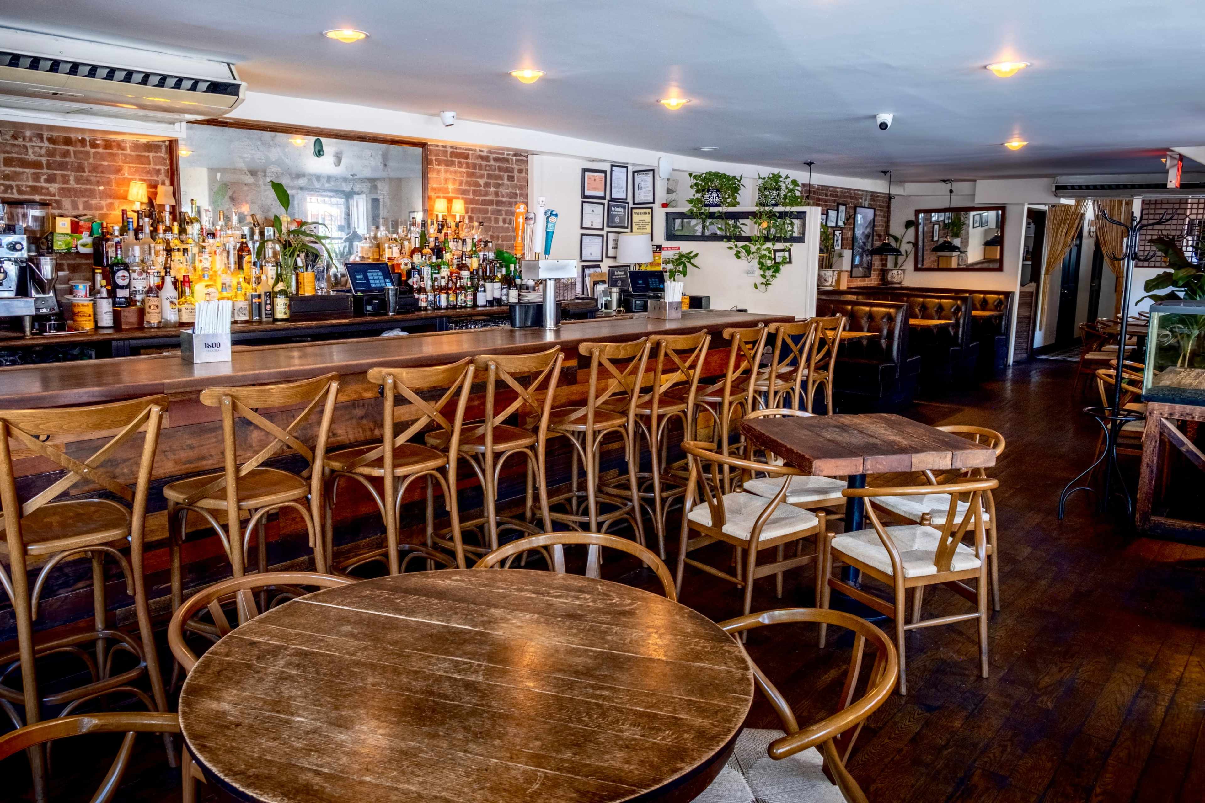 The image shows an interior view of a bar with a long wooden counter, several high-backed chairs, and tables arranged throughout the room.