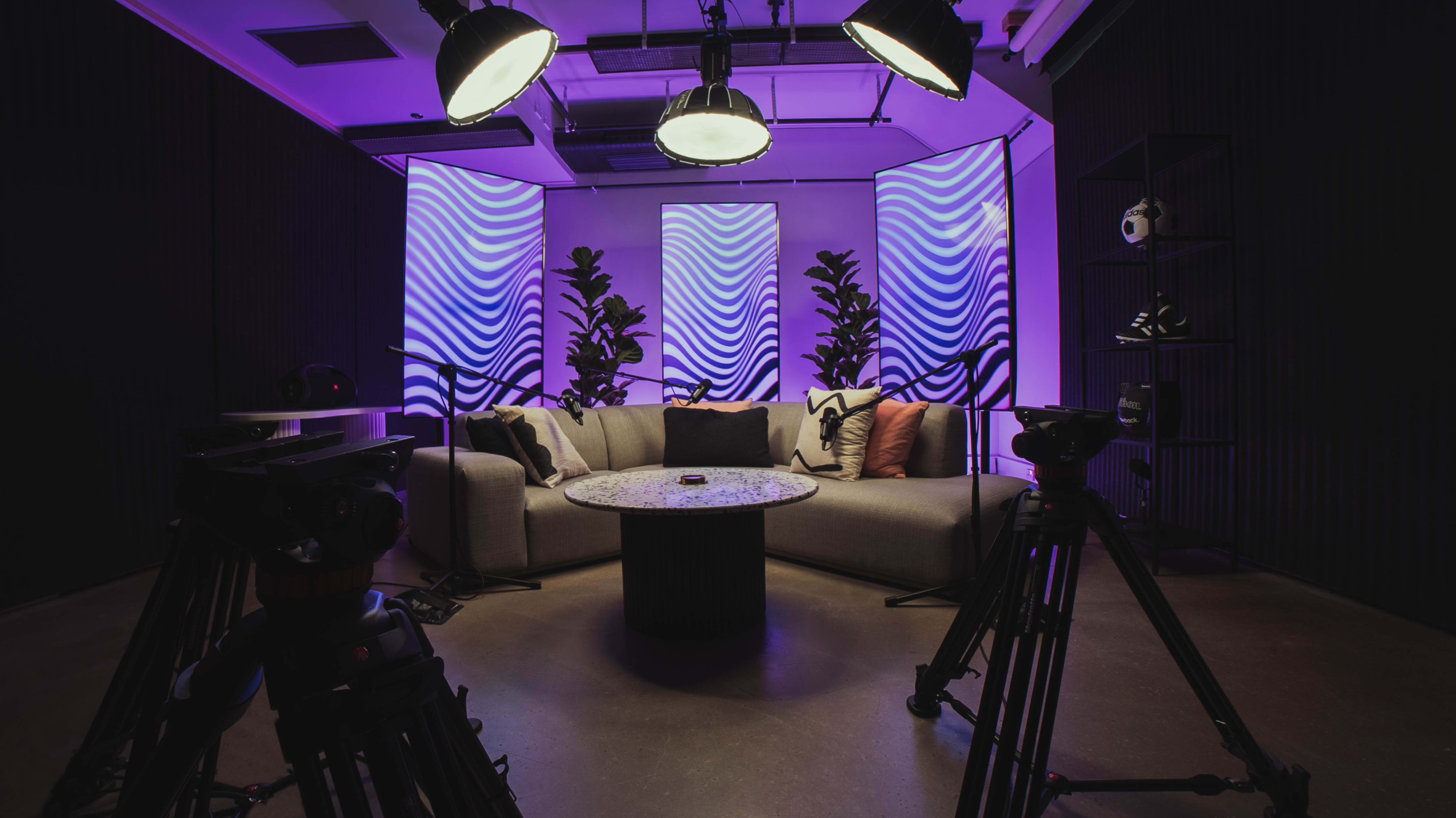 A modern studio setting features a gray couch, a round coffee table, and vibrant purple wave-patterned backdrop elements, with camera equipment positioned in the foreground.