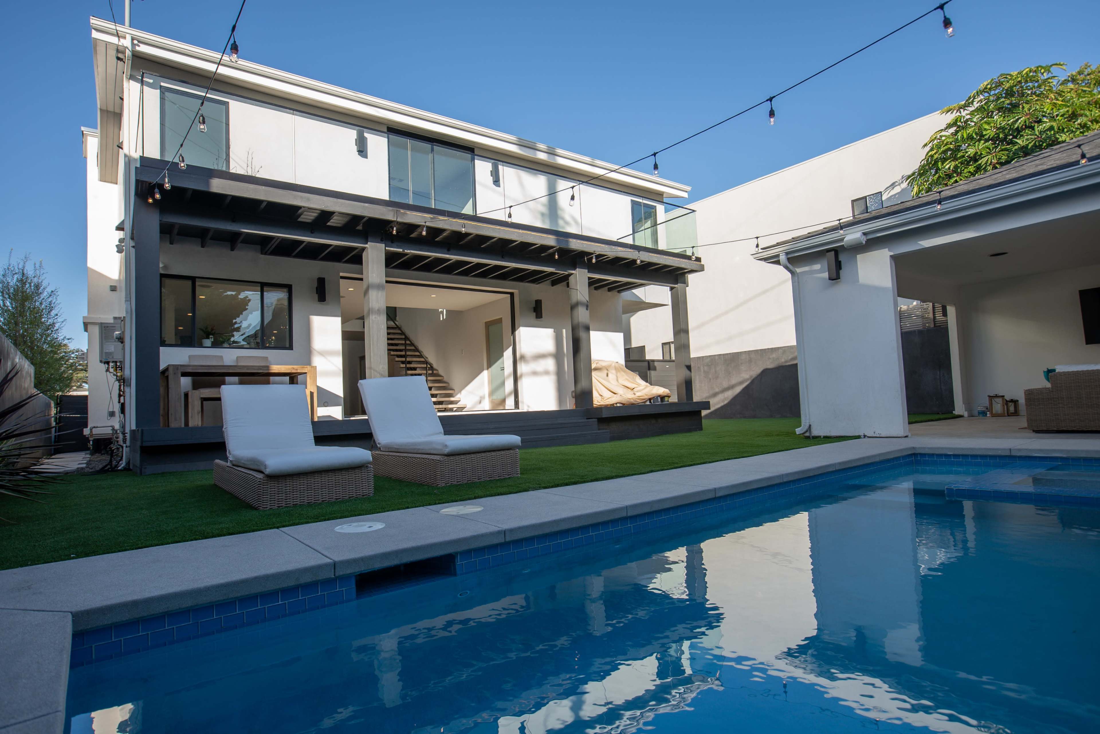 A modern two-story house with a pool, outdoor seating, and stairs leading to a covered patio is set against a clear blue sky.