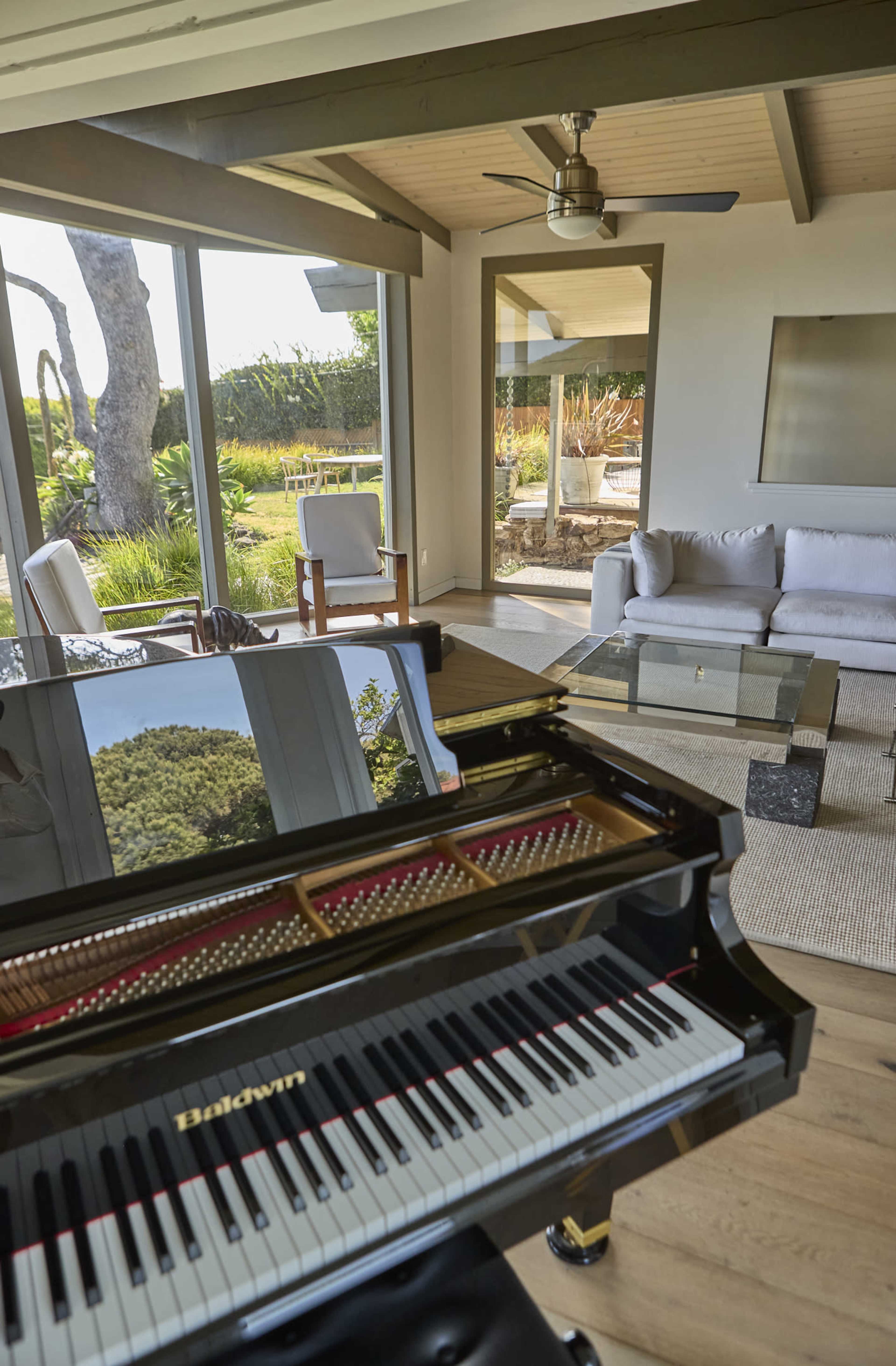 A grand piano sits in the foreground of a modern living space that features large glass windows overlooking a landscaped garden.