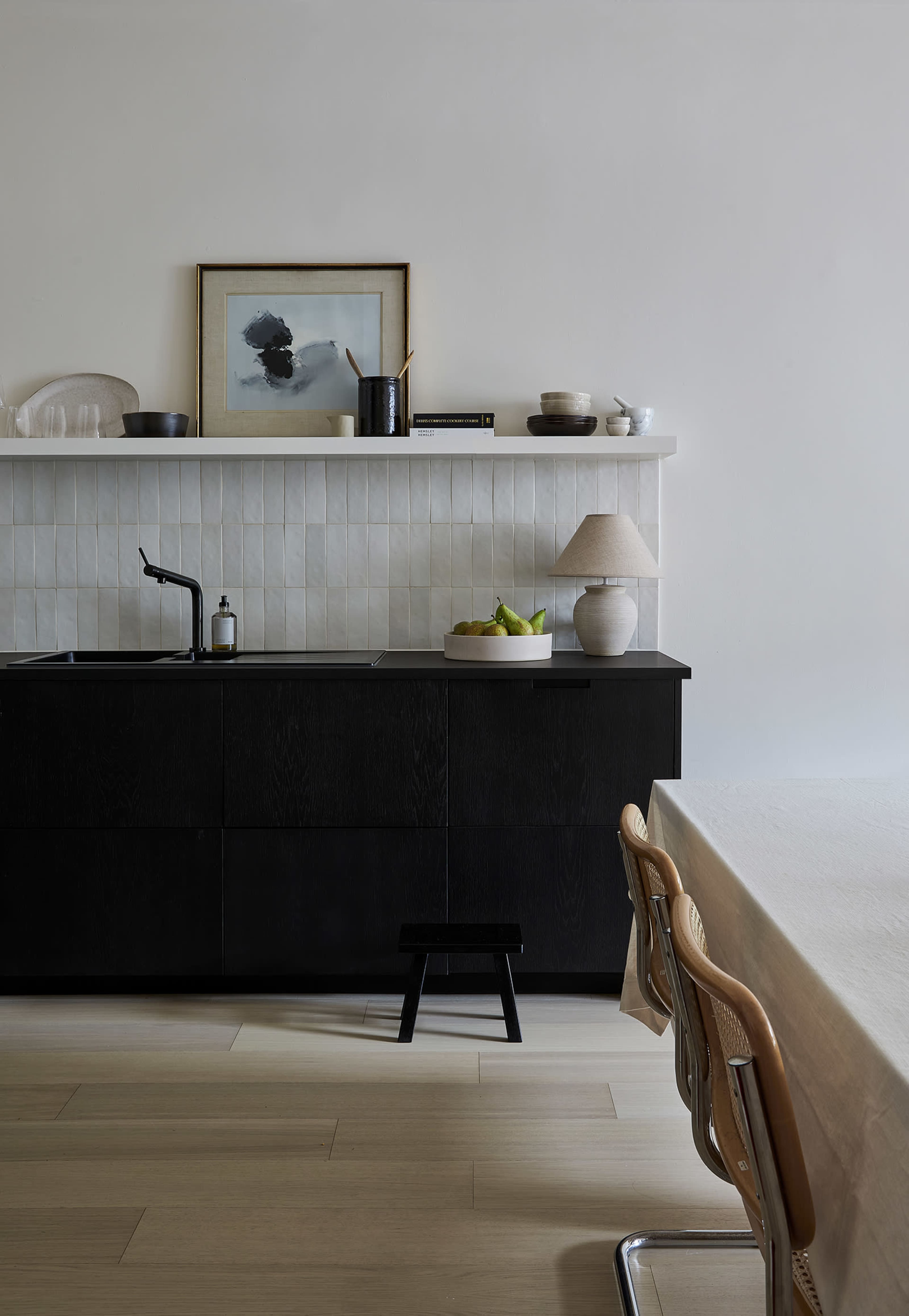 A minimalist kitchen features a black cabinet, a white countertop with decorative items, and a light wooden dining table with metal-framed chairs.