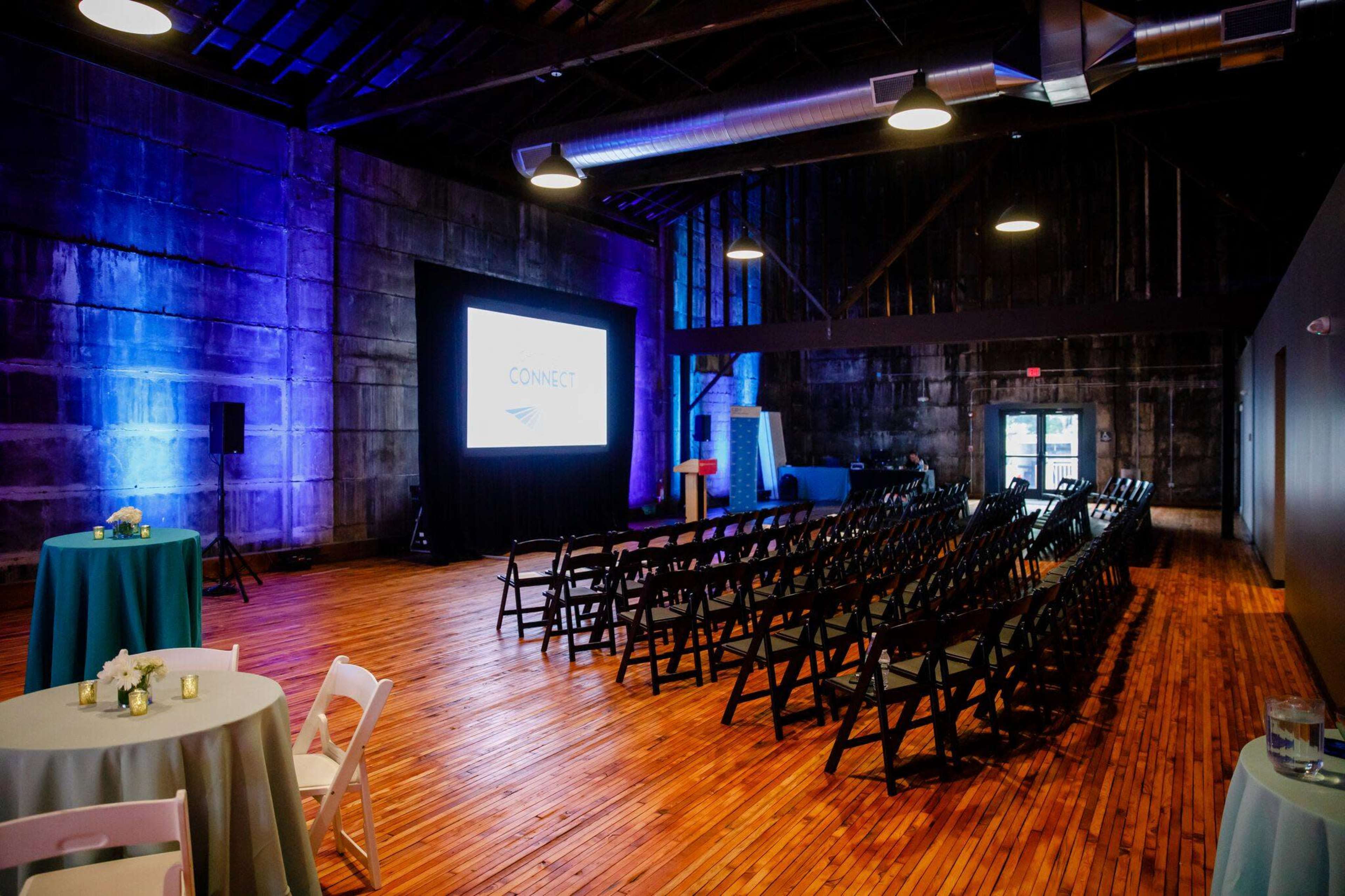 A dimly lit event space with wooden flooring, featuring rows of black chairs facing a screen, surrounded by round tables.