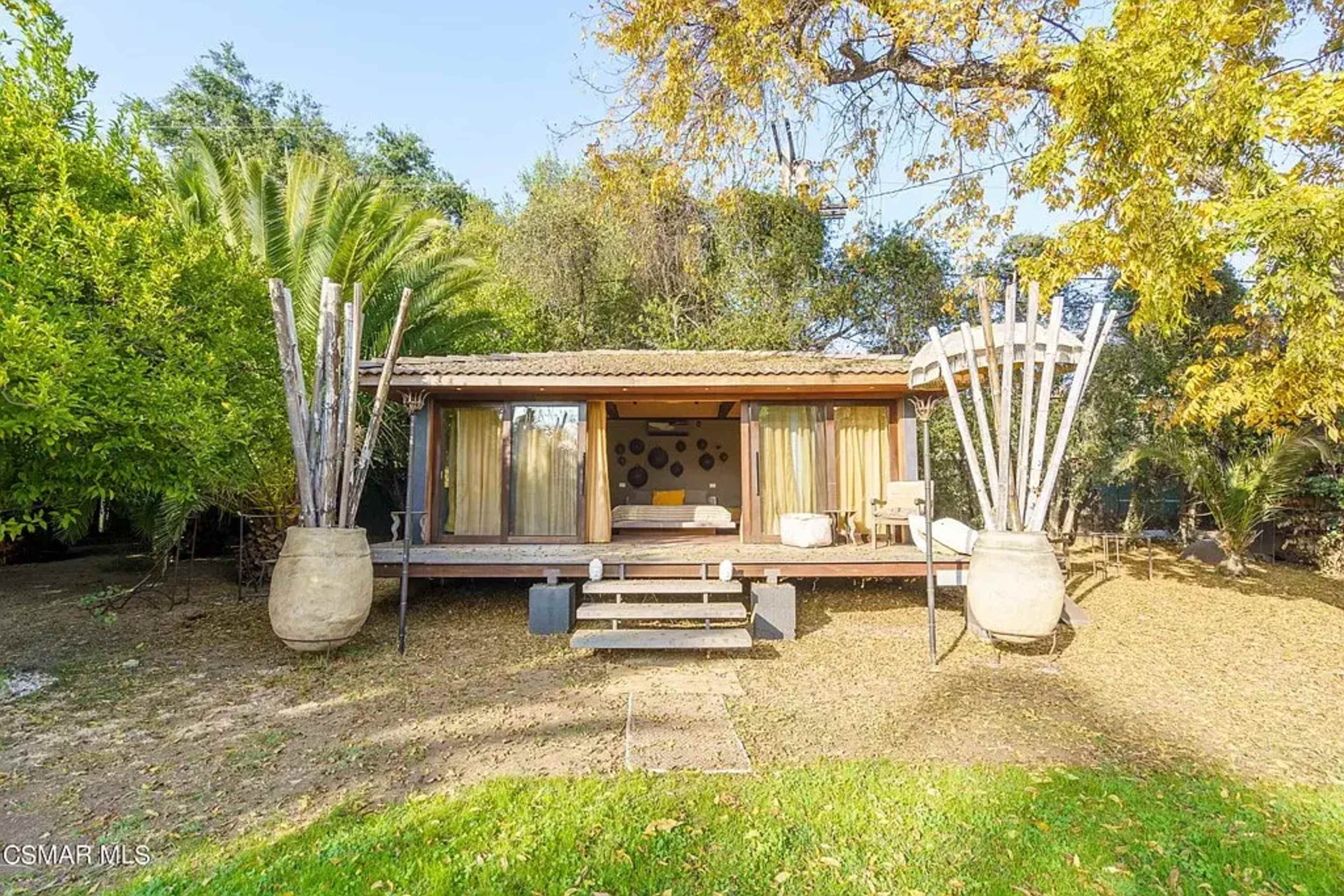 A wooden cabin with large windows is surrounded by greenery and features two large decorative pots at the entrance.
