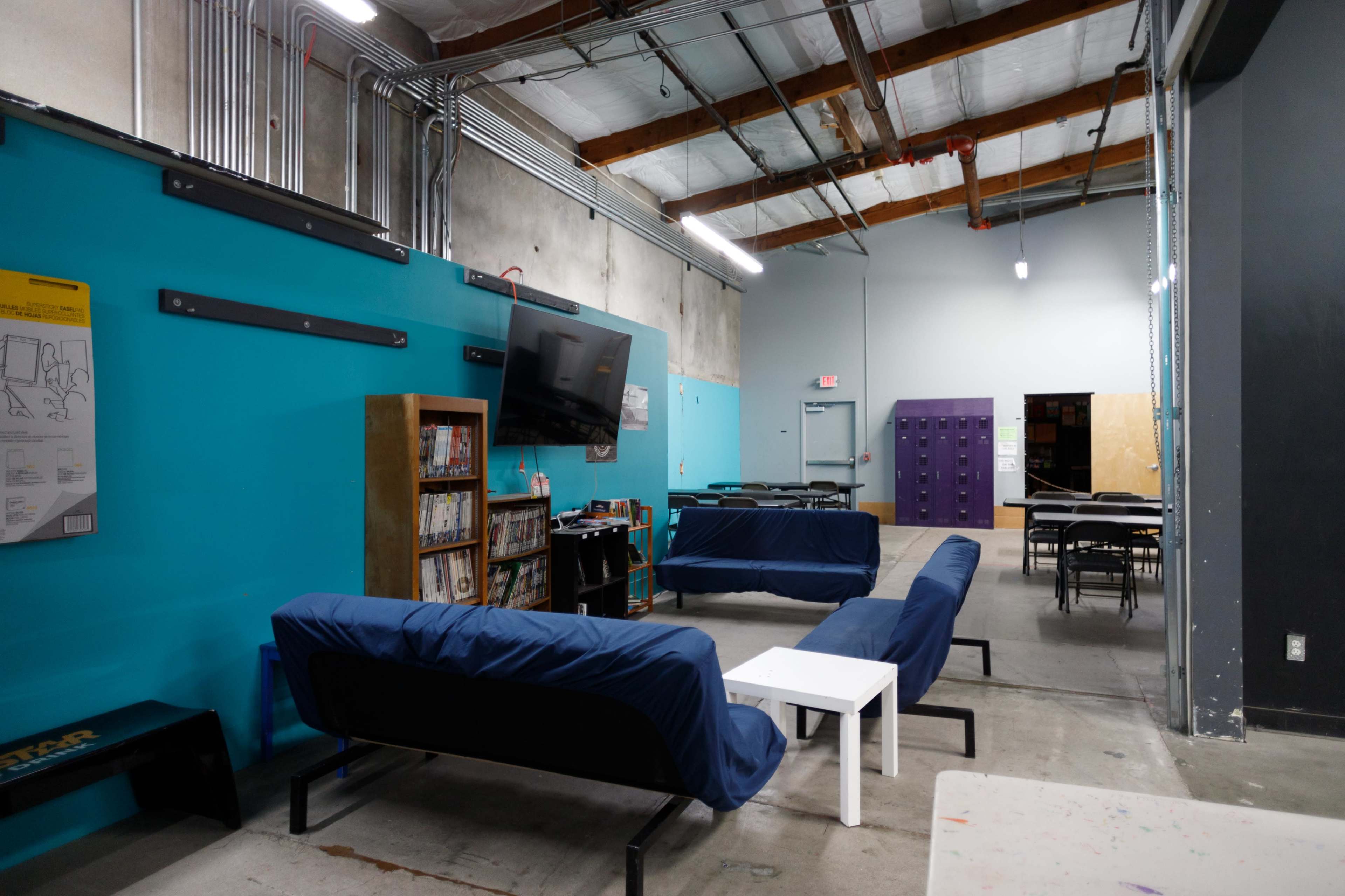 The image shows a spacious room with blue walls, featuring two couches, a white table, a bookshelf, a television, and purple lockers in the background.
