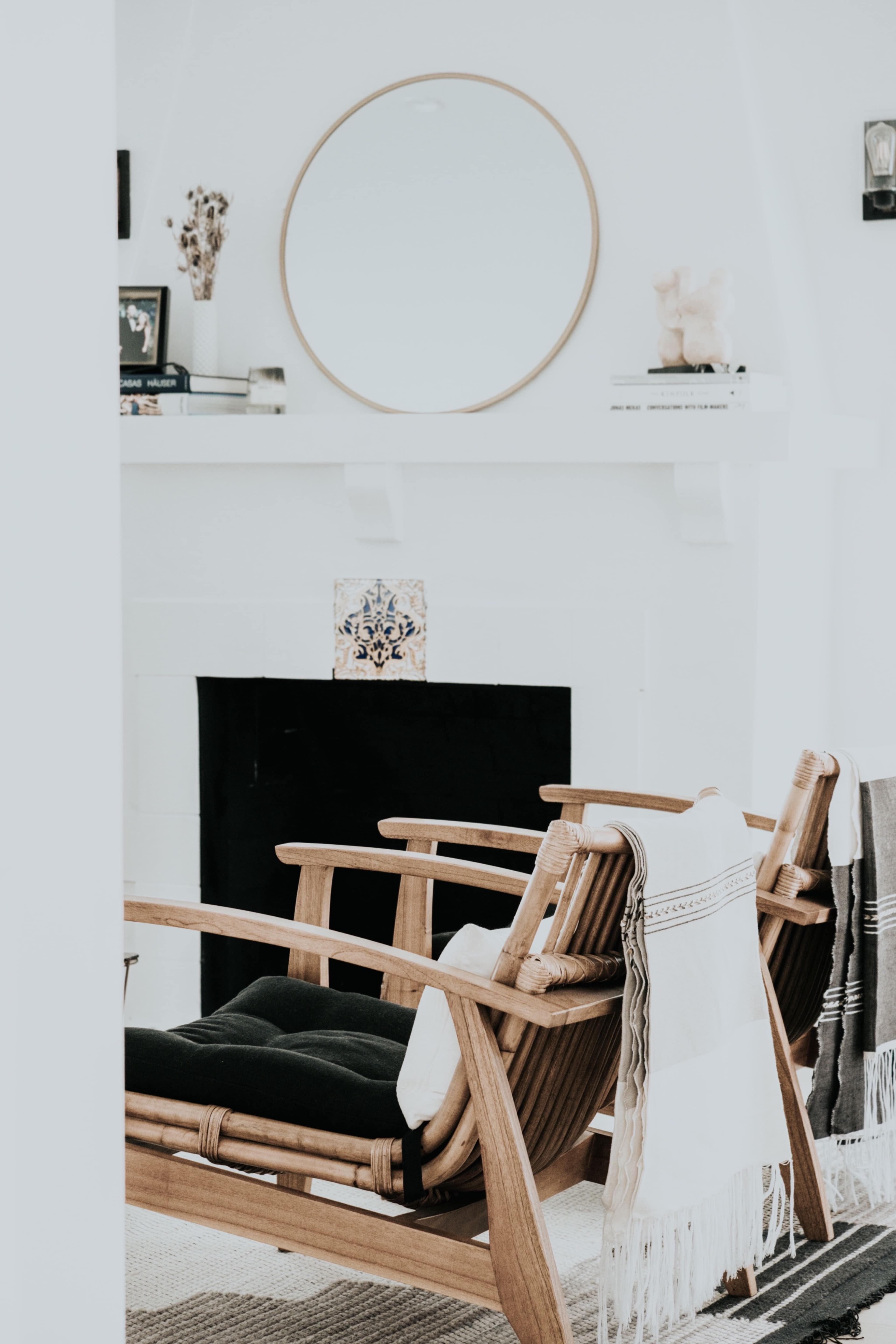 The image shows a cozy living room with two wooden chairs, a circular mirror above a white fireplace, and decorative items on the mantle.