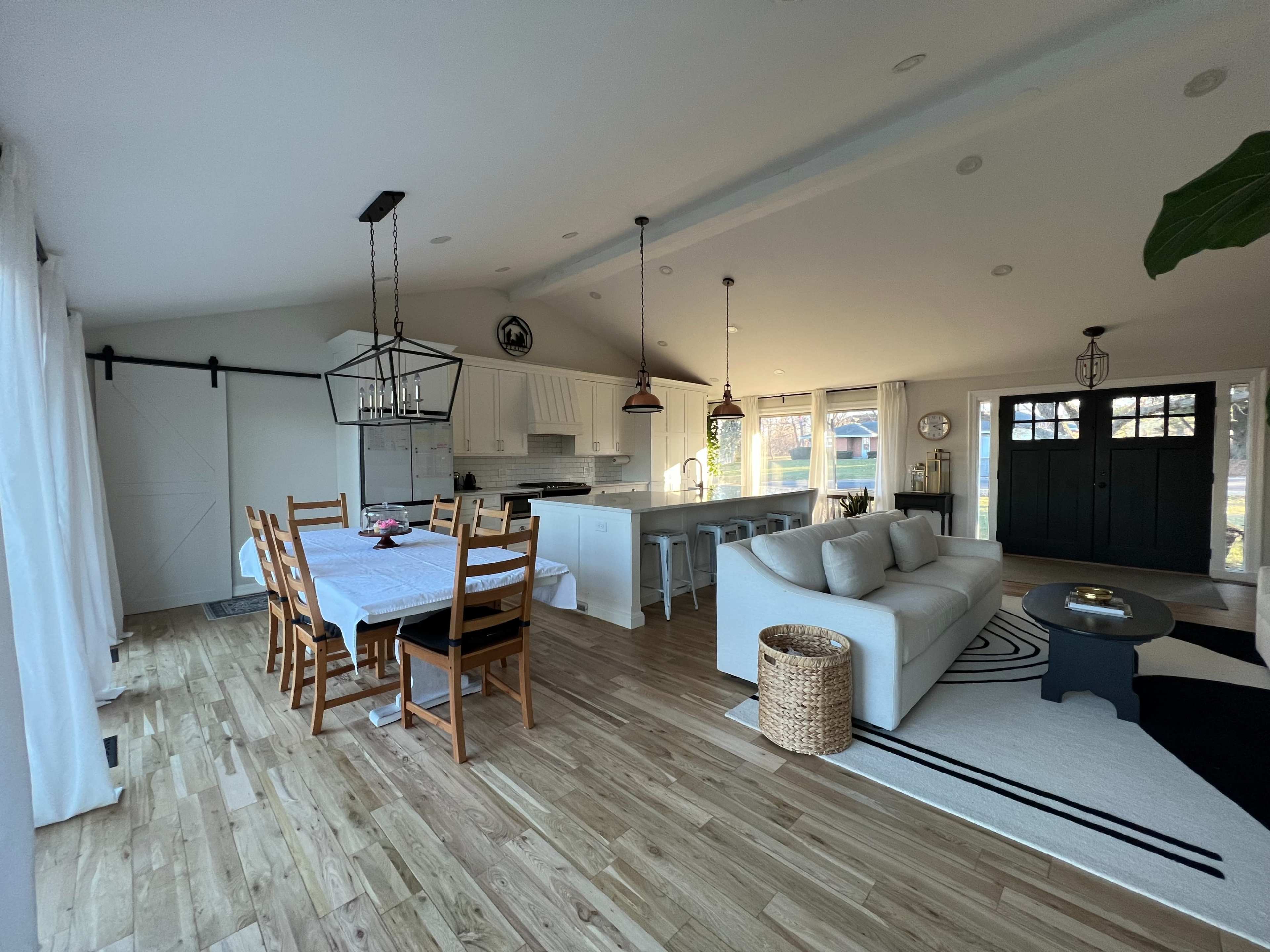 A modern, open-concept dining and living area with a wooden table, chairs, a sofa, and a kitchen in the background.
