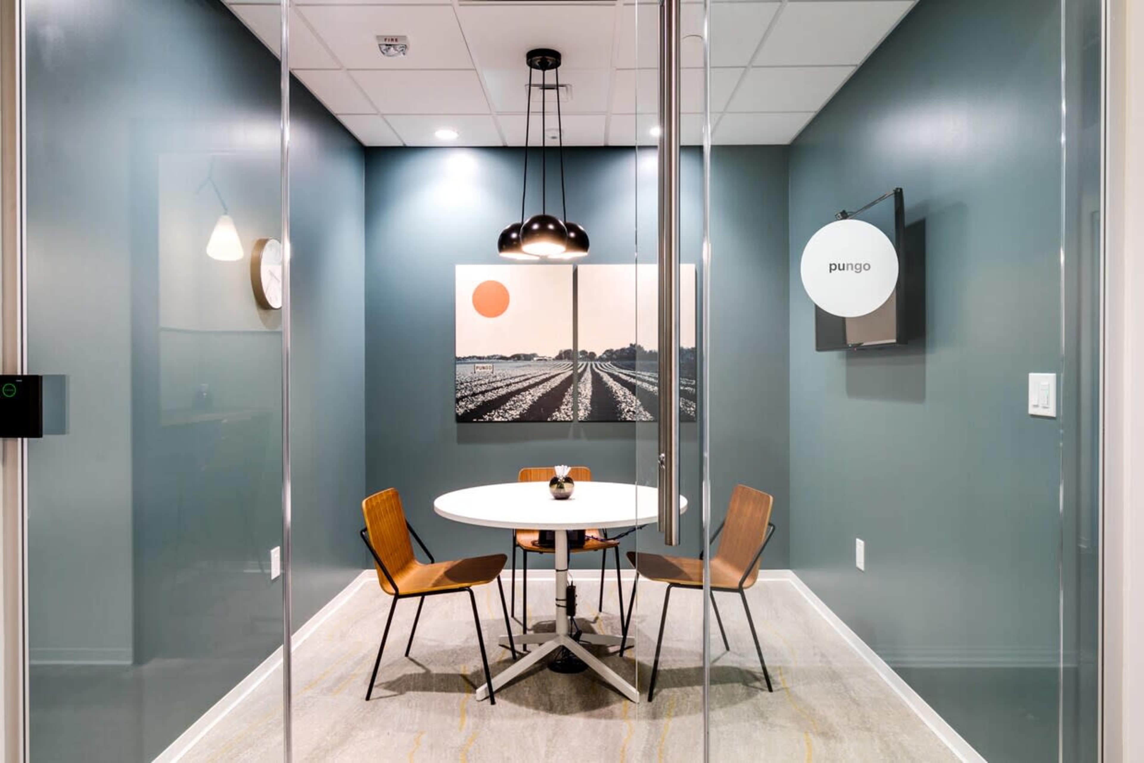 A small conference room features a round table with three wooden chairs, a large artwork on the wall, and a modern light fixture hanging from the ceiling.
