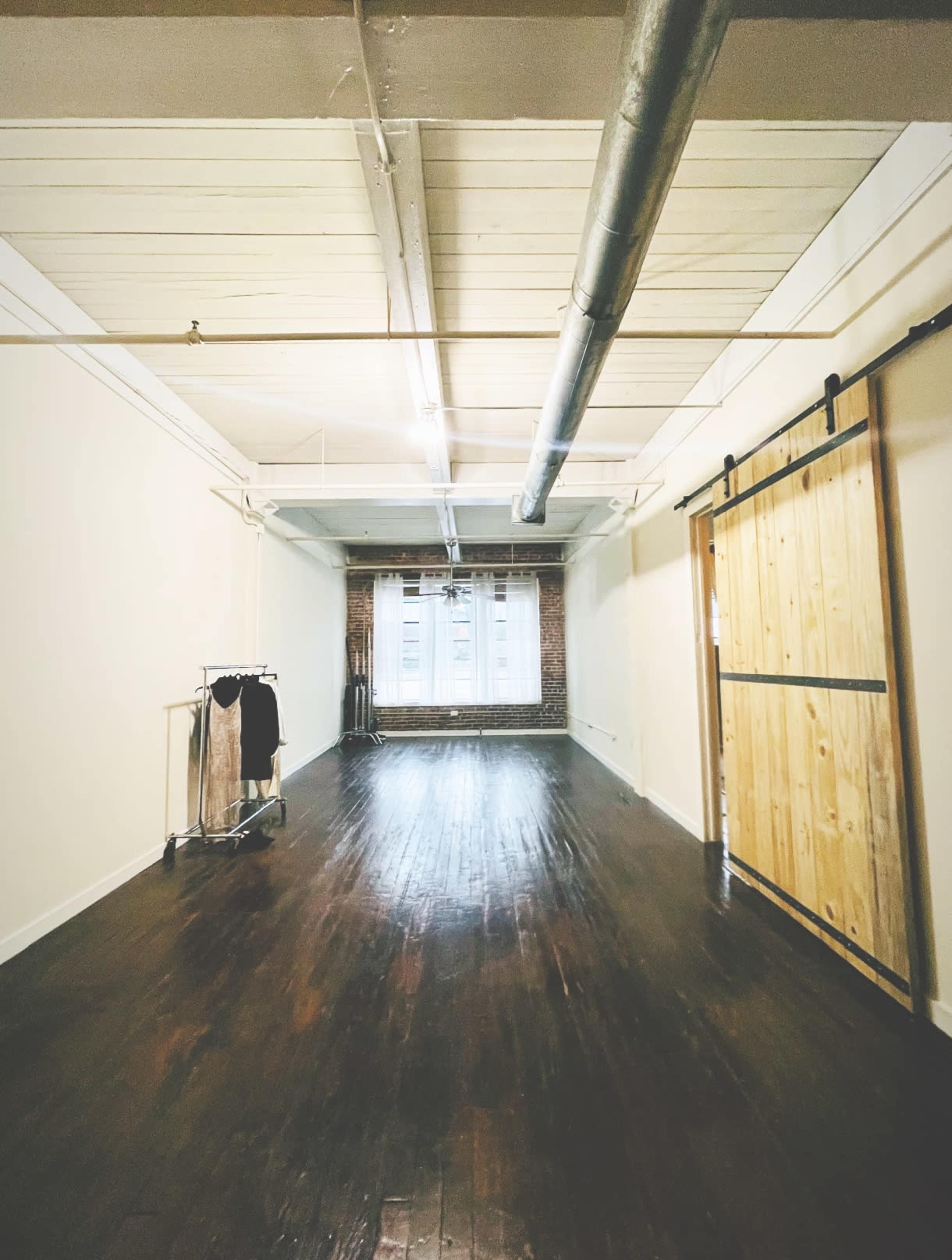 A spacious room features wooden floors, exposed brick walls, and a large window with sheer curtains.
