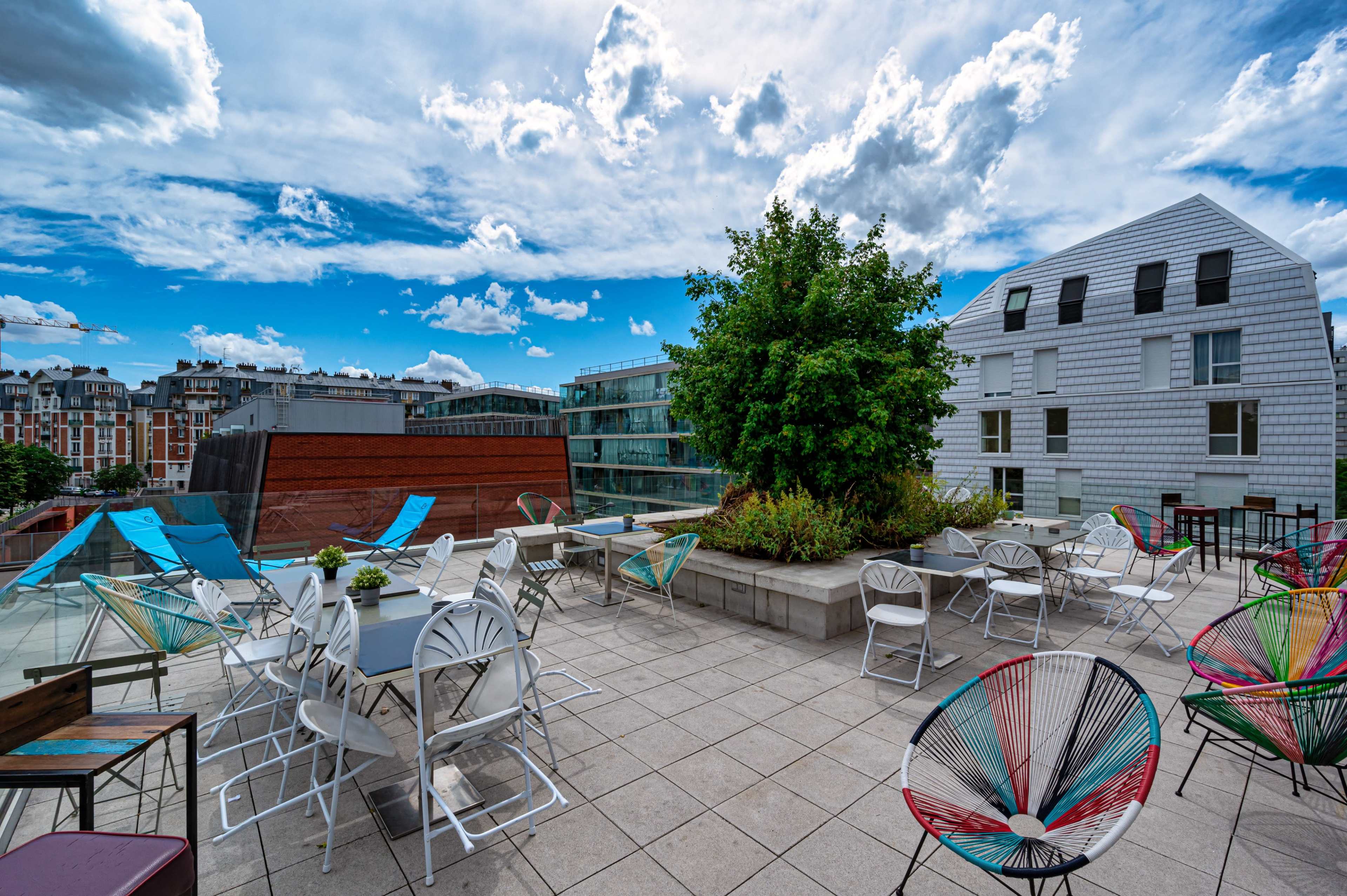 The rooftop terrace features a variety of tables and chairs surrounded by greenery under a cloudy sky.