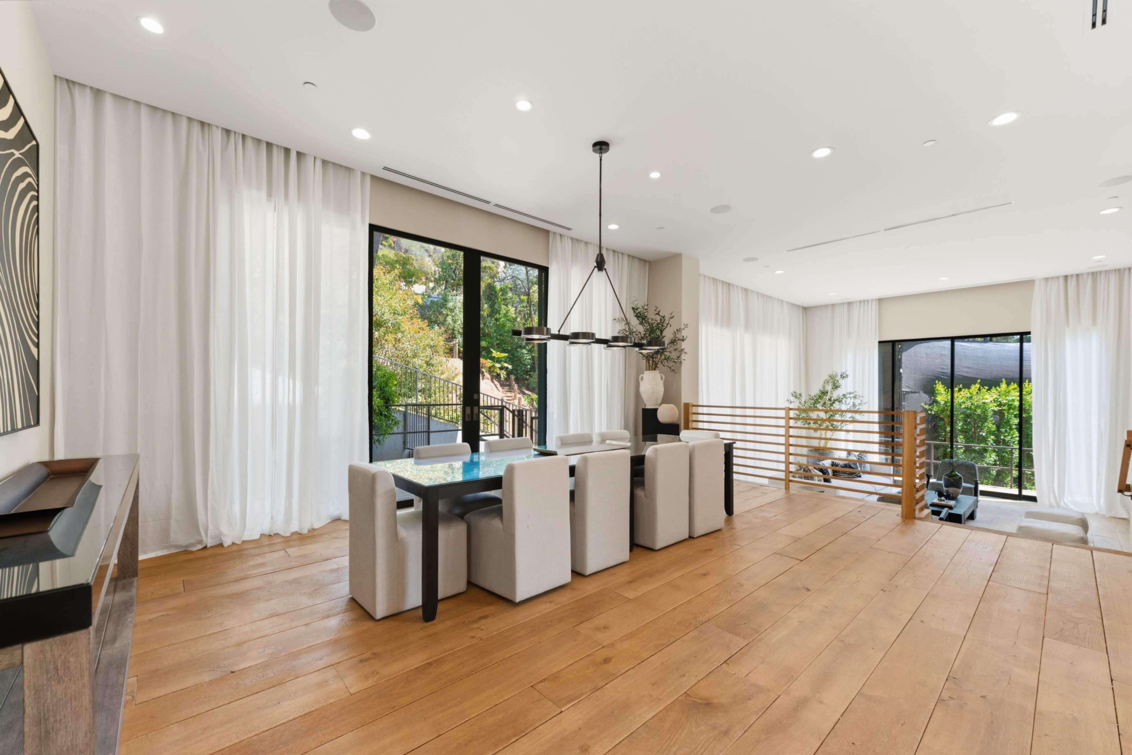 The image shows a modern, open-concept dining area with a wooden floor, a large dining table surrounded by upholstered chairs, and large windows letting in natural light.