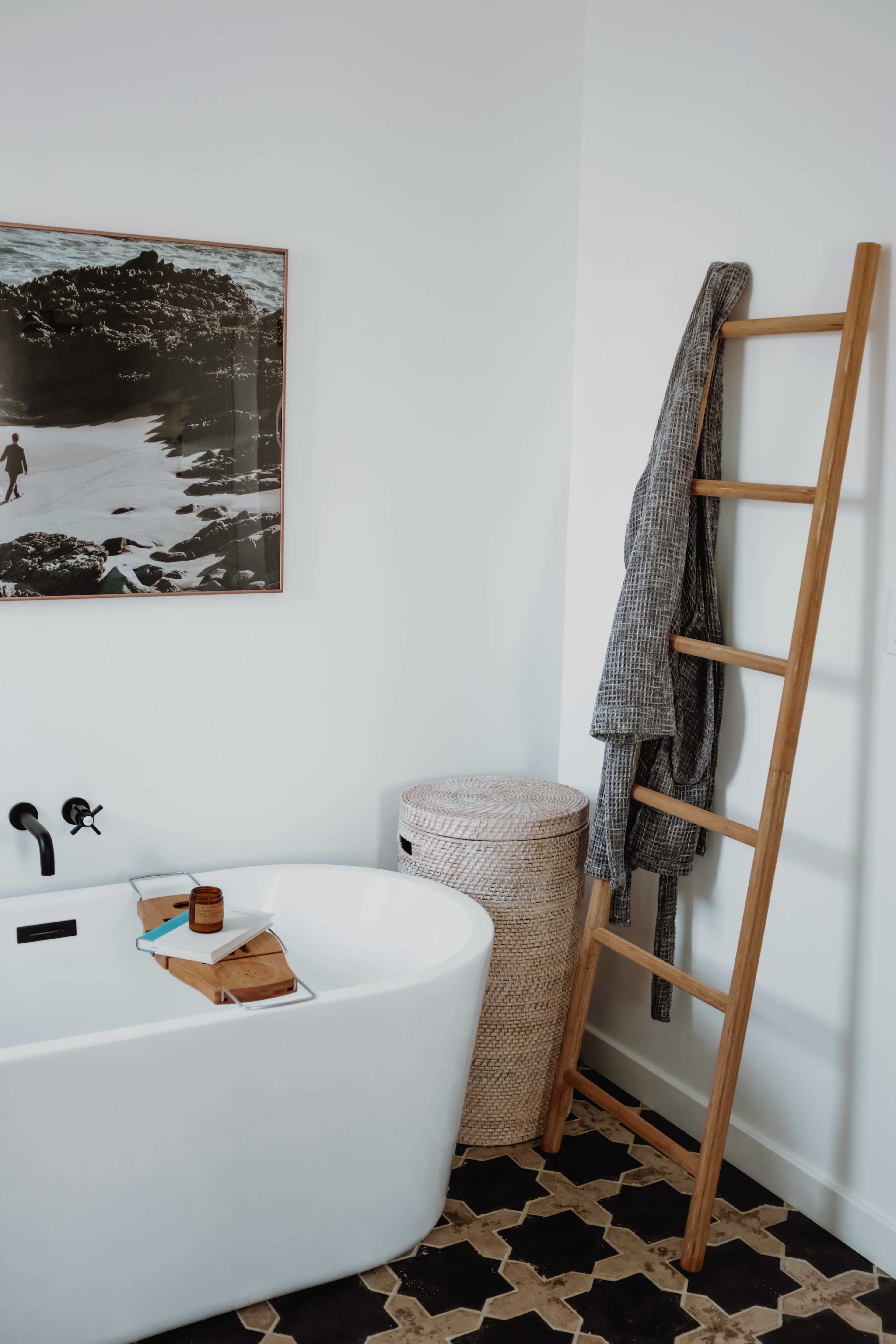 The image features a modern bathroom with a freestanding white tub, a woven laundry basket, and a wooden ladder holding a robe, complemented by a black-and-white photograph on the wall.