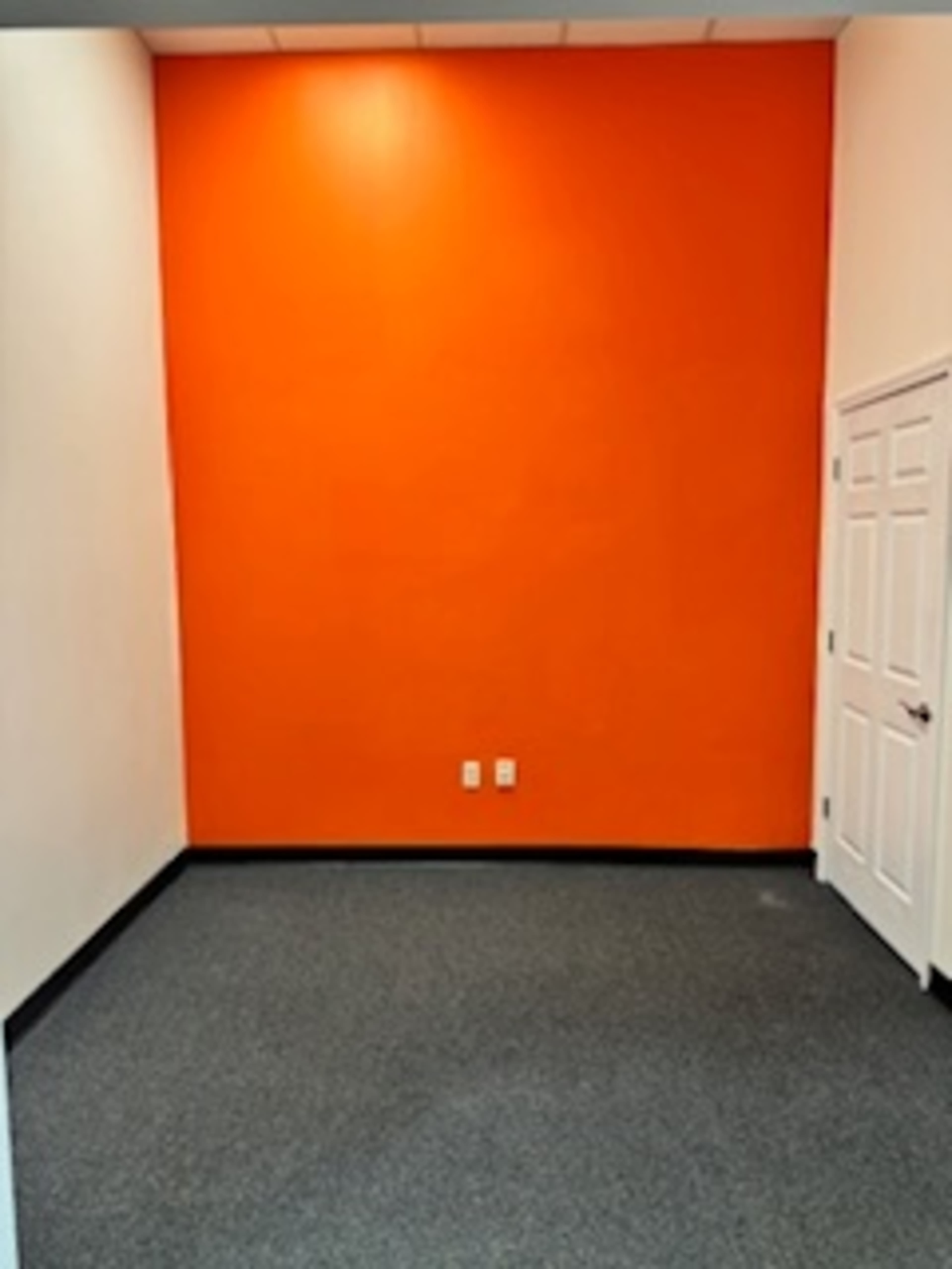 The image depicts a small room with an orange accent wall, gray carpeted flooring, and a white door on one side.