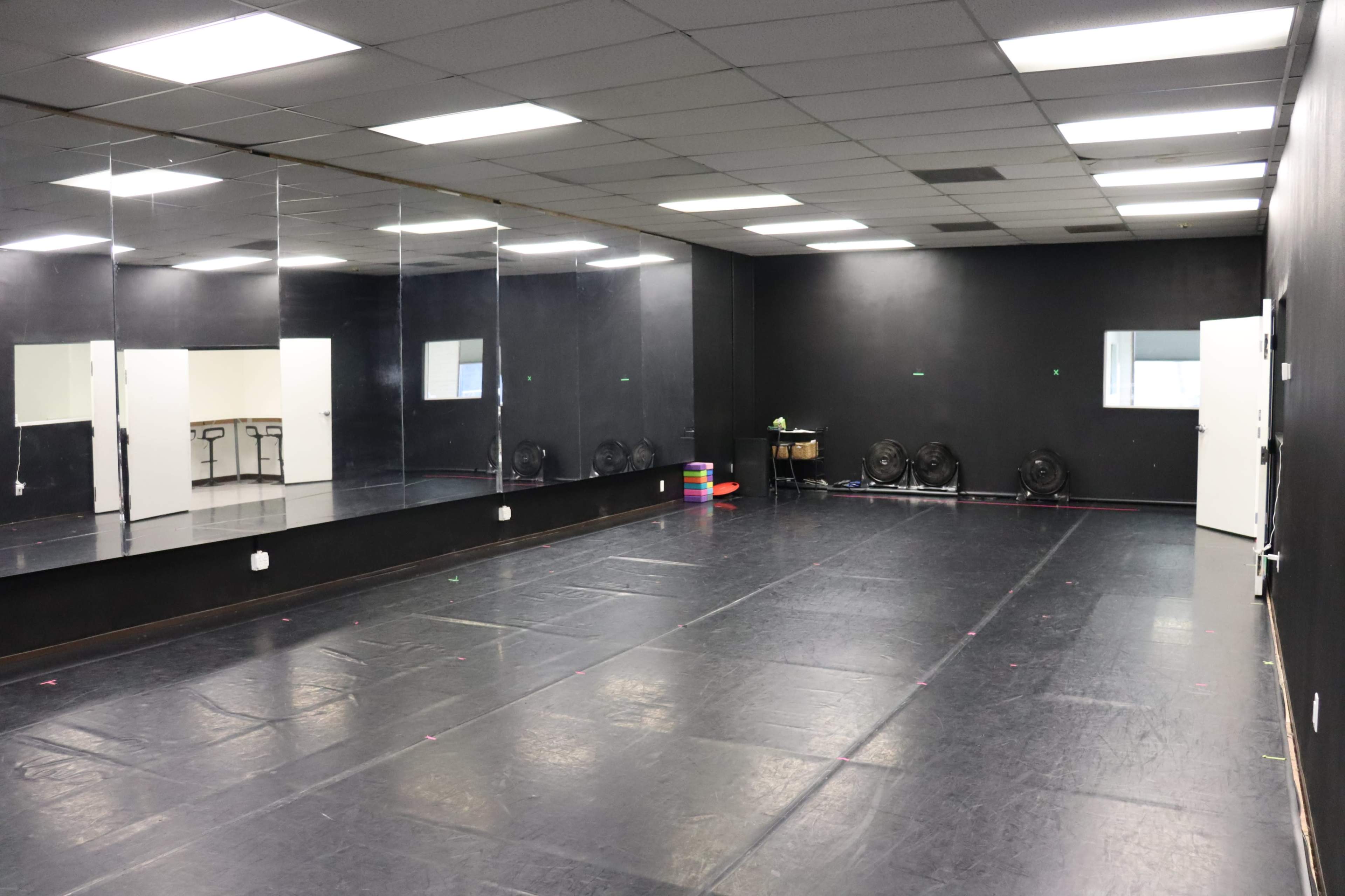 The image shows an empty dance studio with a black floor, mirrors on one wall, and several fans positioned around the room.