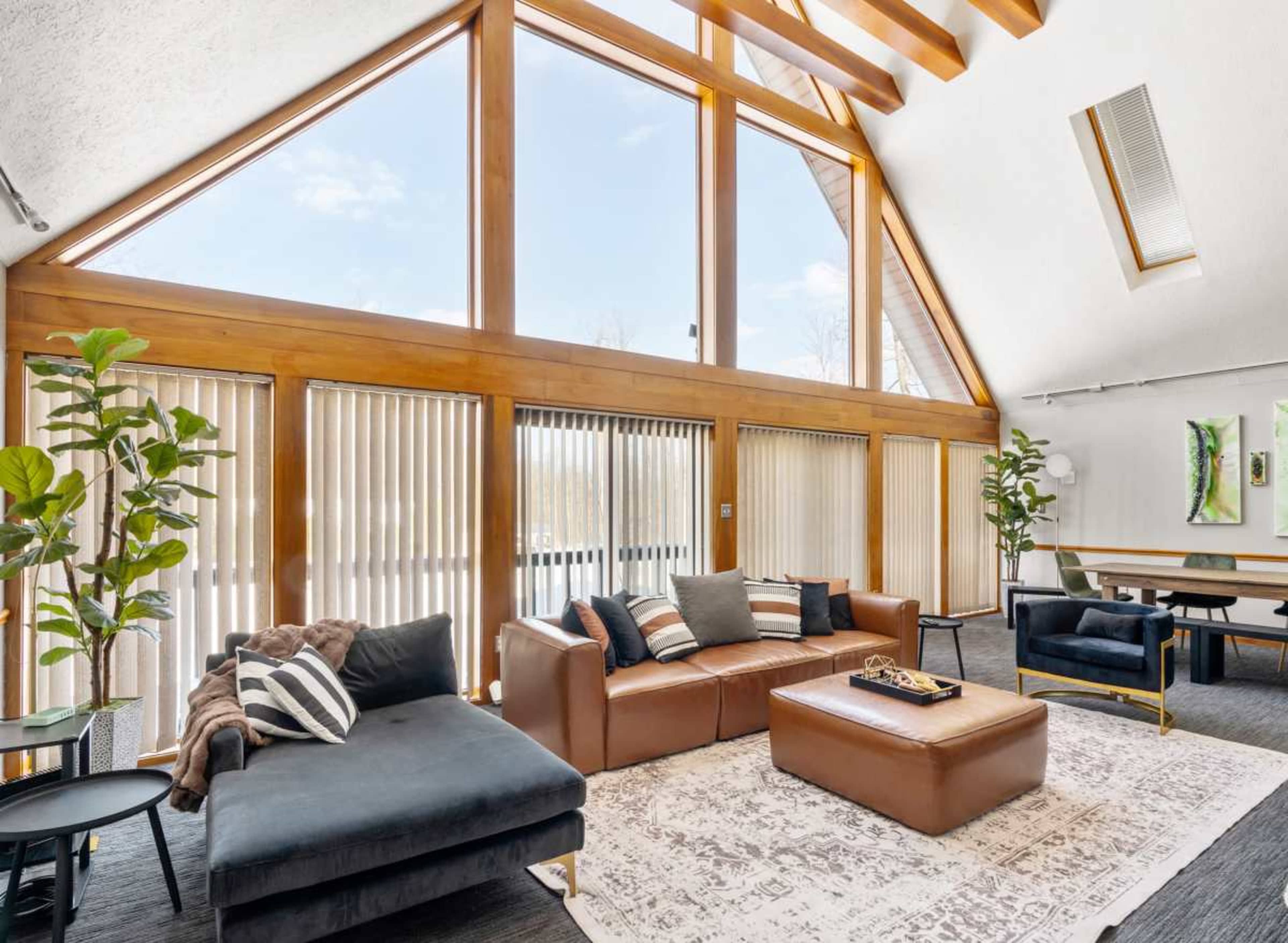 The image shows a spacious living area with large windows, wooden beams, and modern furniture including a sectional sofa and coffee table.