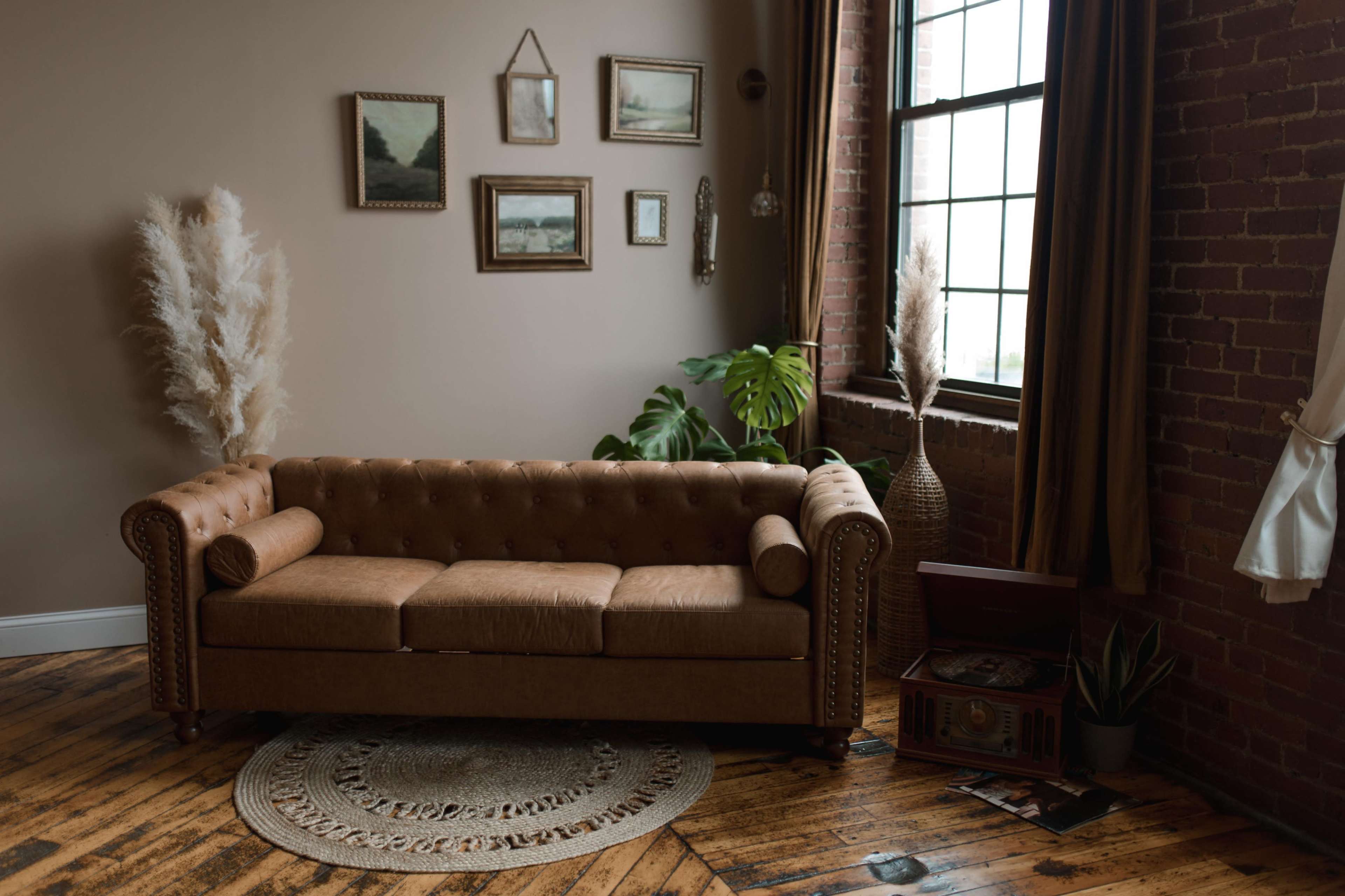 The image shows a brown tufted sofa in a room with brick walls, decorated with framed pictures and plants.