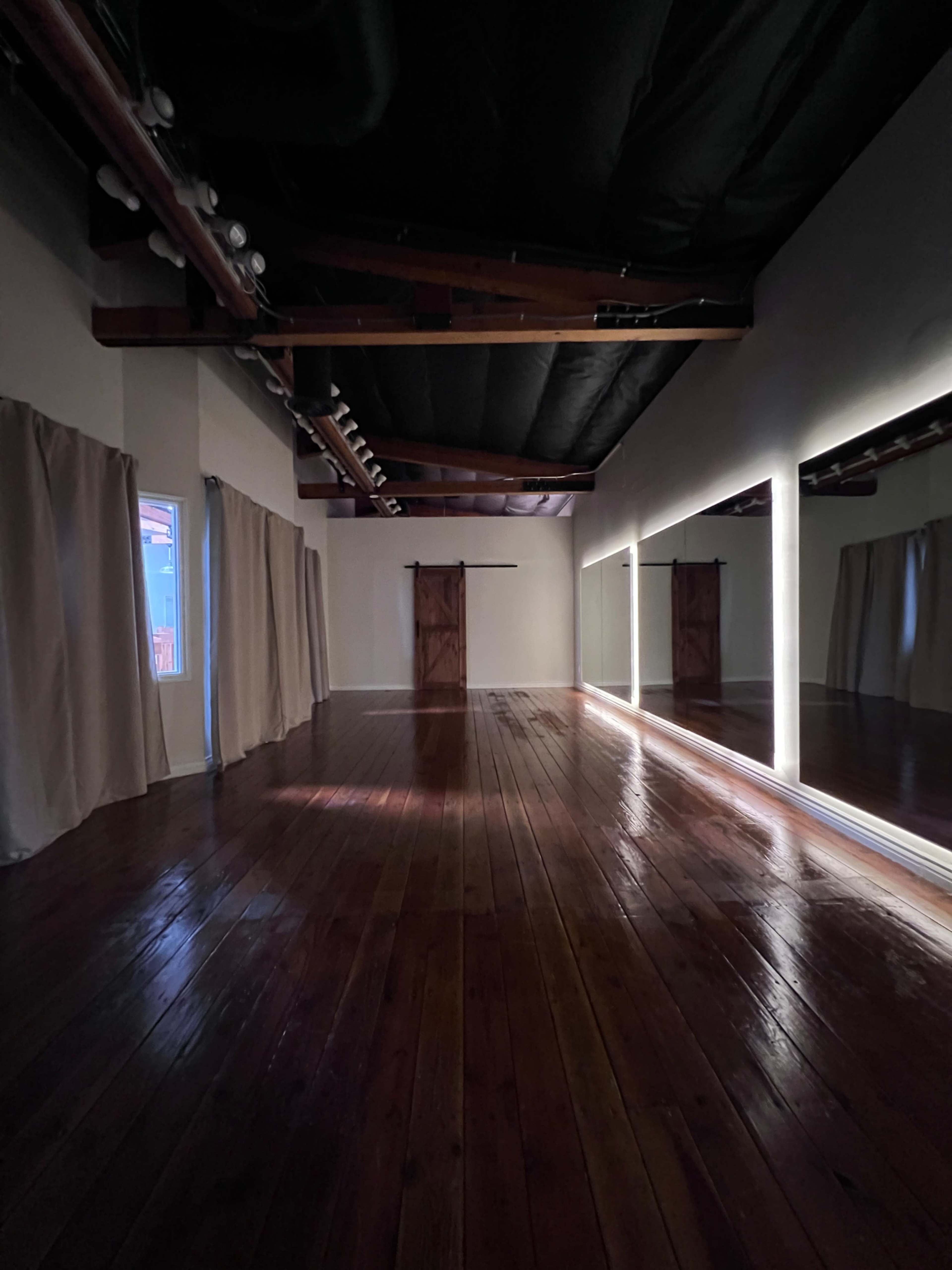 The image shows an empty room with wooden floors, large windows covered by curtains, and a long mirror illuminated by a light strip along one wall.