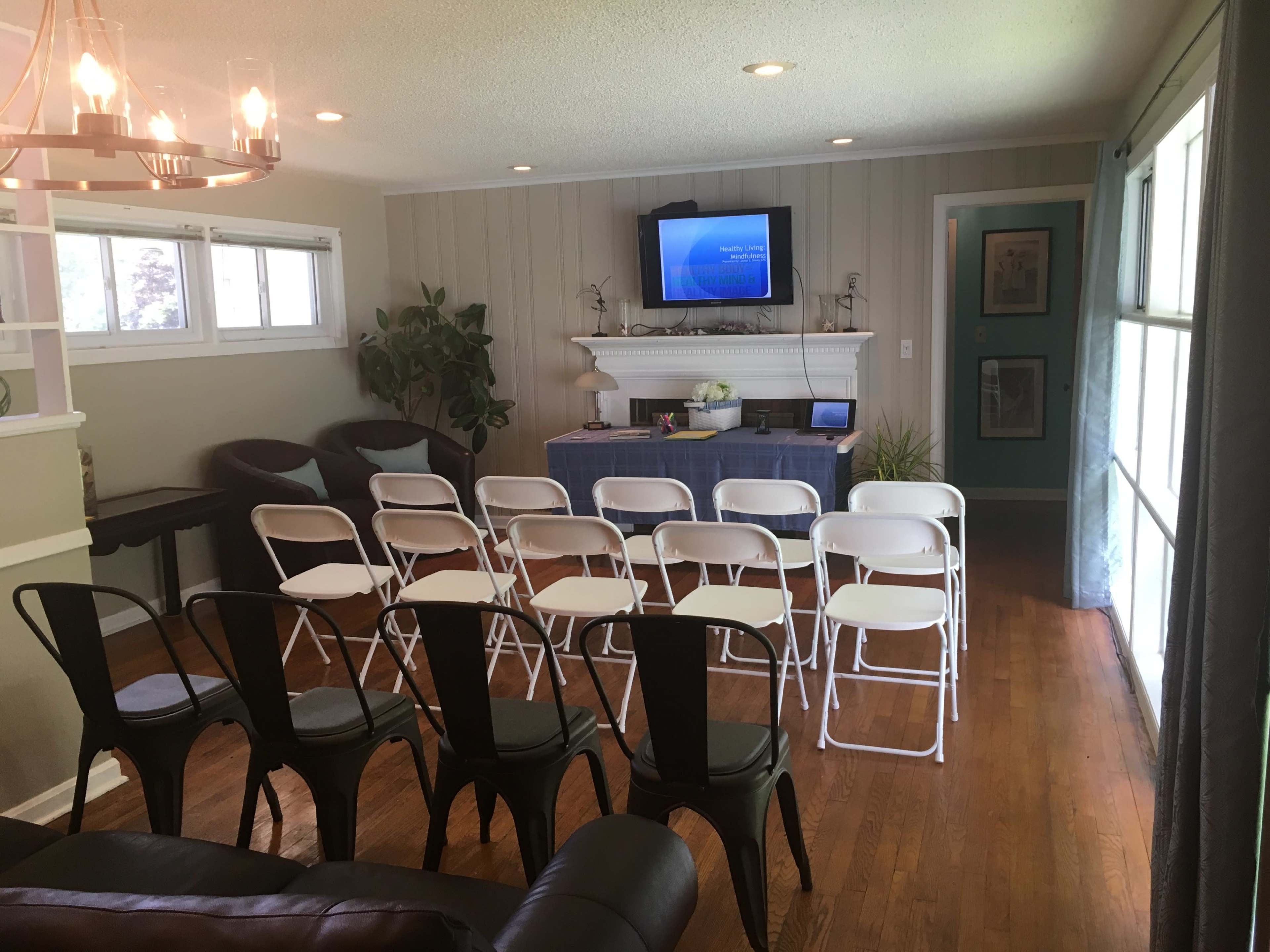 A room set up for a presentation, featuring several rows of folding chairs facing a screen on the wall, with a table in front displaying a laptop and plants.
