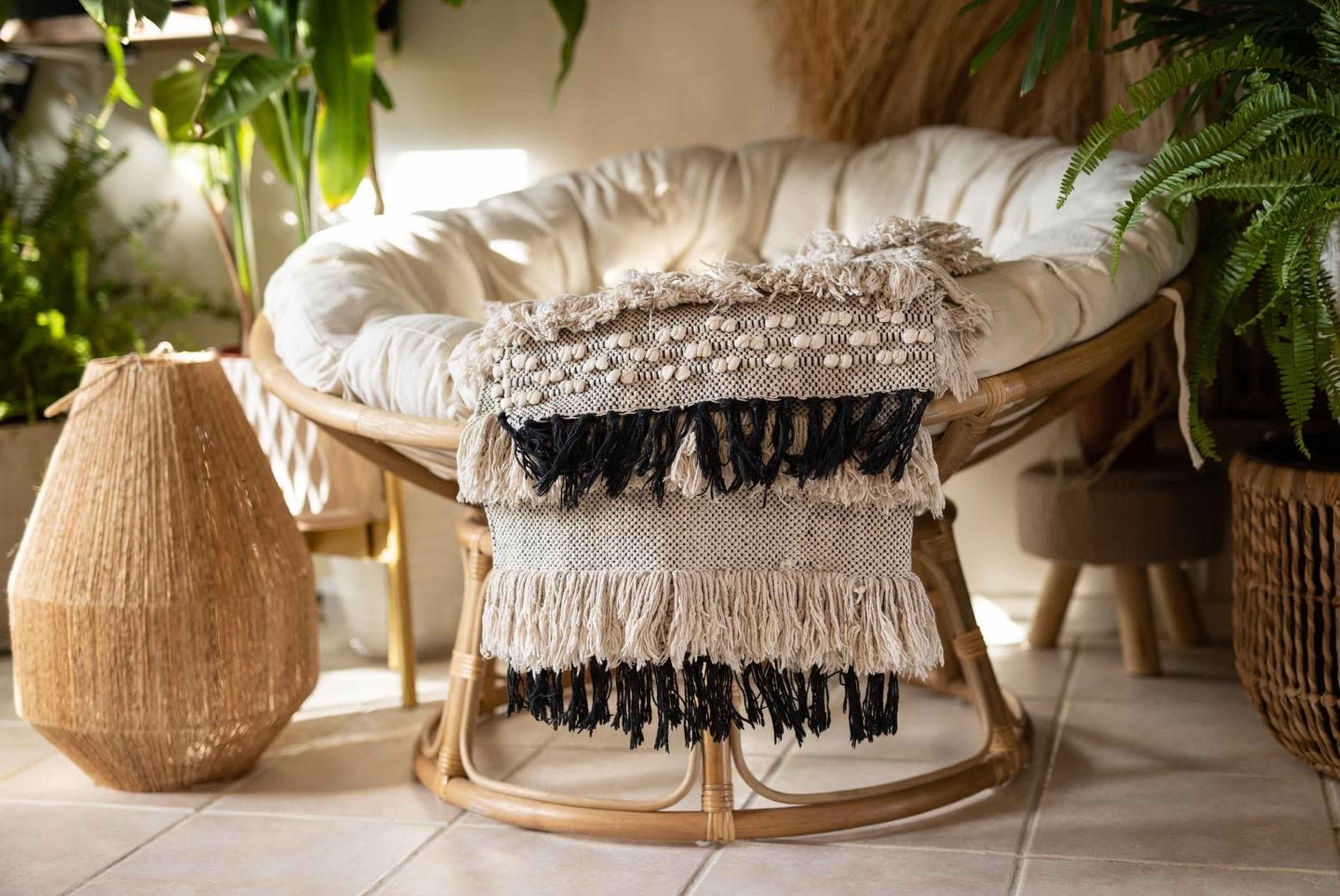 A woven chair with a cushion is draped with a textured blanket, surrounded by various indoor plants and natural decor elements.