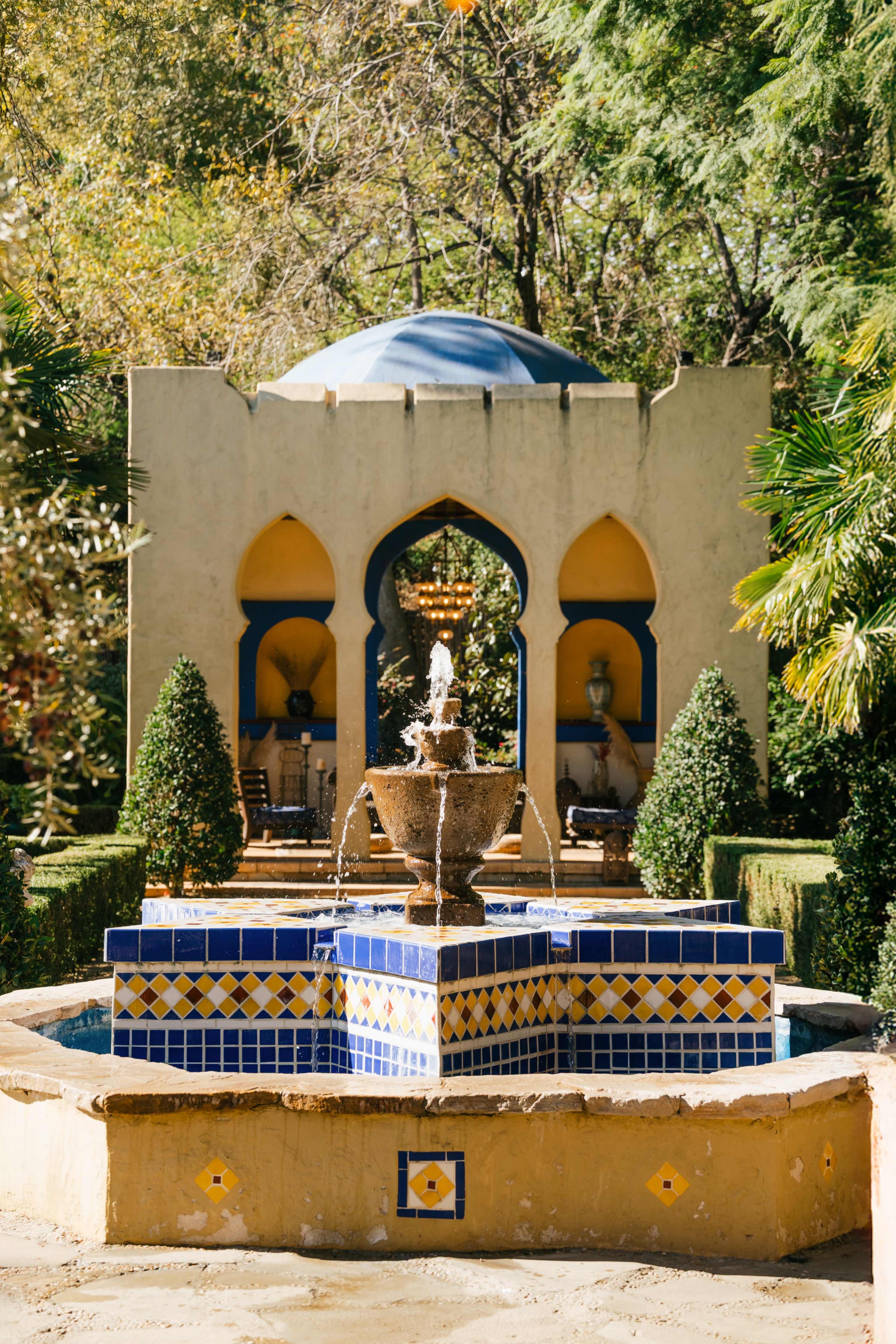 The image shows a decorative garden with a central fountain surrounded by tiled seating and lush greenery, framed by an architectural structure featuring arches and a domed roof.