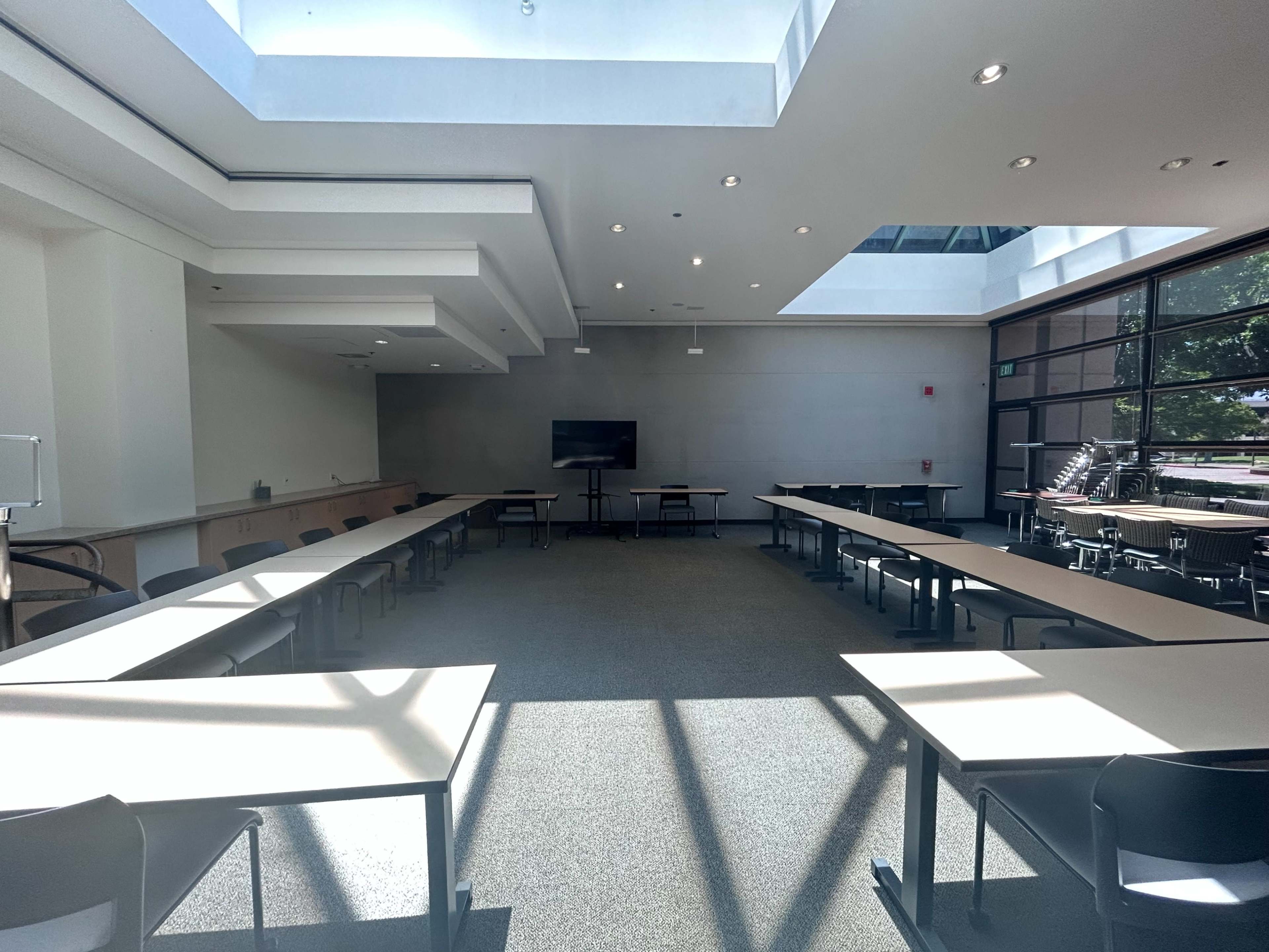 A well-lit meeting room features several tables arranged in a U-shape and a large screen at the front.